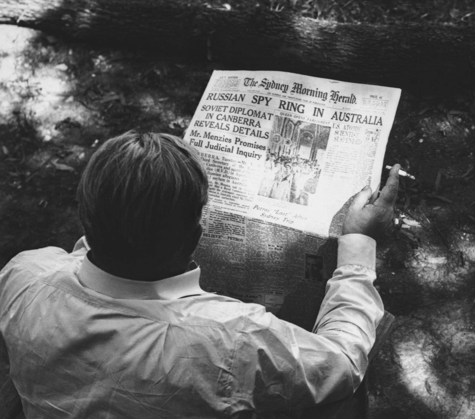 A man in a shirt and holding a cigarette in his right hand is shown from behind holding a newspaper - a copy of the Sydney Morning Herald. The front page is visible over this shoulder and the headline says "Russian spy ring in Australia'.