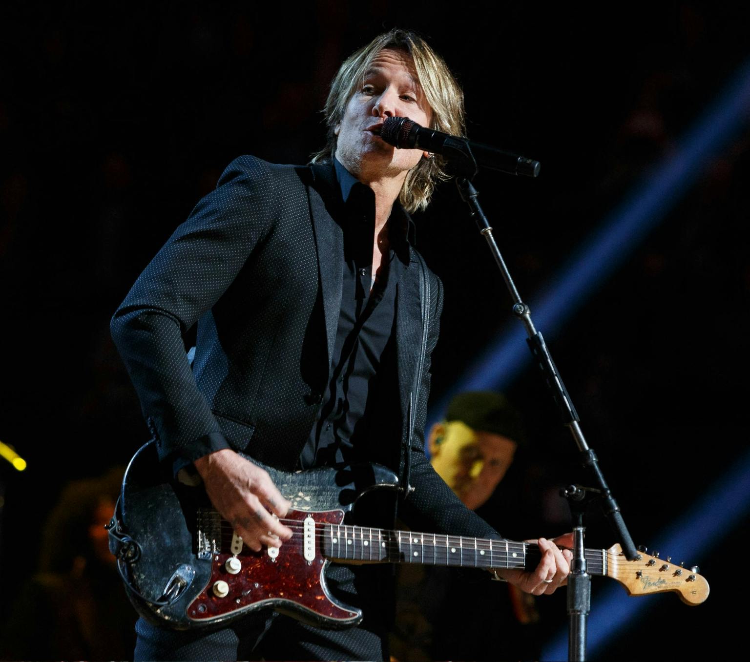 A man with medium length brown hair is playing a guitar while singing into a microphone. he is wearing a black suit and black shirt