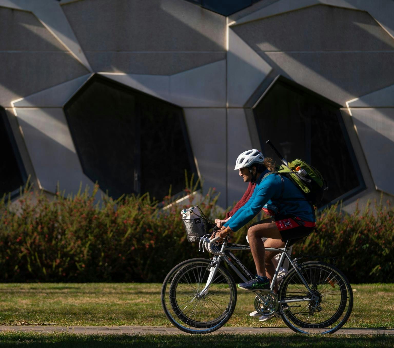 Cyclist rides a bike along a path in front of a geometric building.