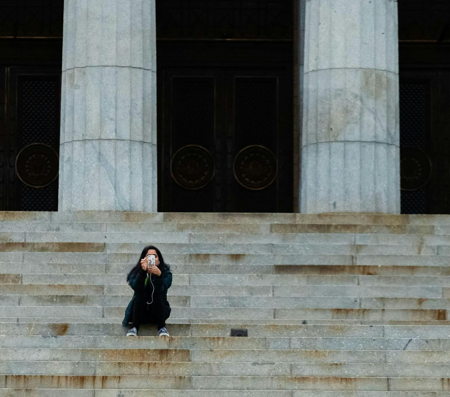 young person looking at their phone and sitting on a set of sandstone steps with large round columns behind. There is no one else in the image