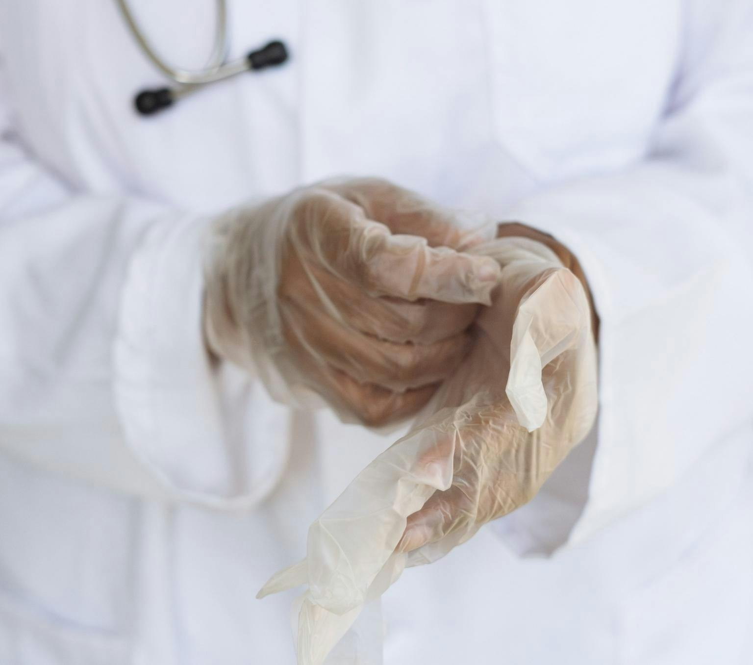Close up of a person's hands putting on clear plastic gloves. The person is wearing a white lab coat and has a stethoscope around their neck