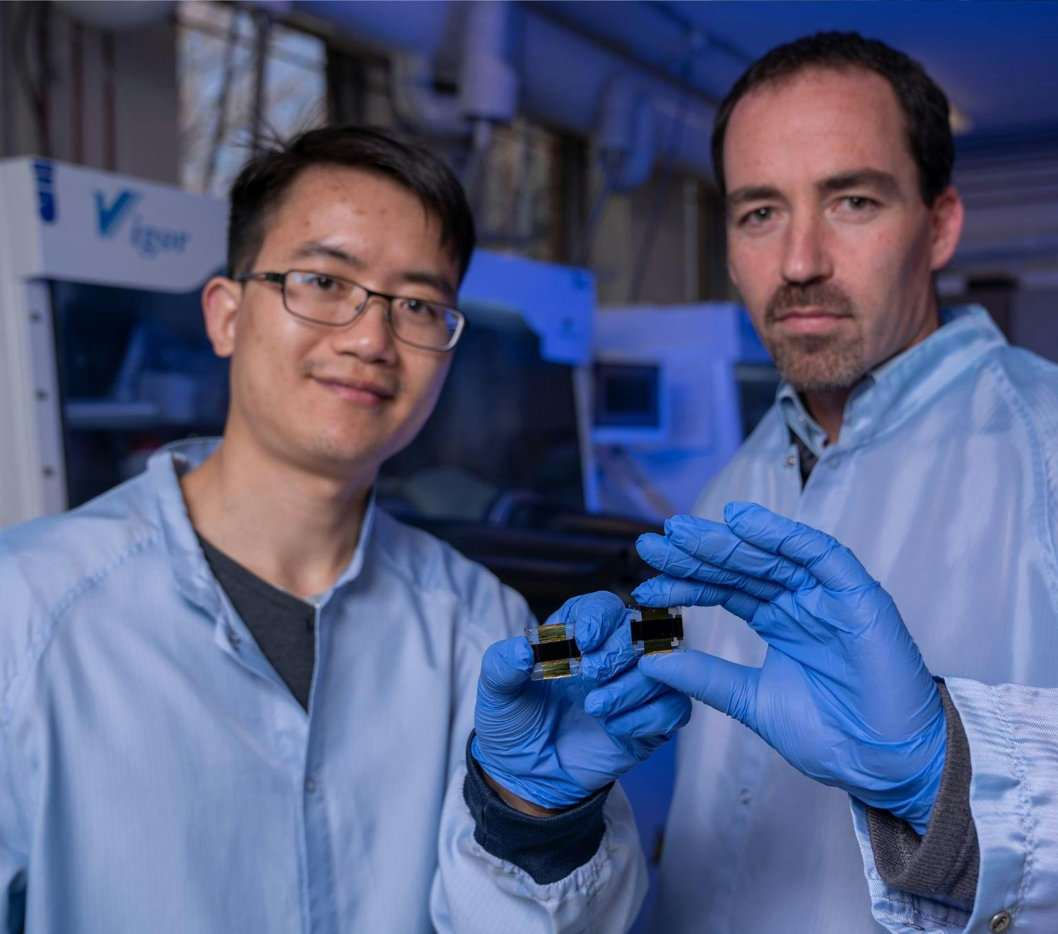 Jun Peng and Thomas White are both wearing blue lab coats and blue golves. ther are holding small gold and black square shaped cells and standing in a laboratory