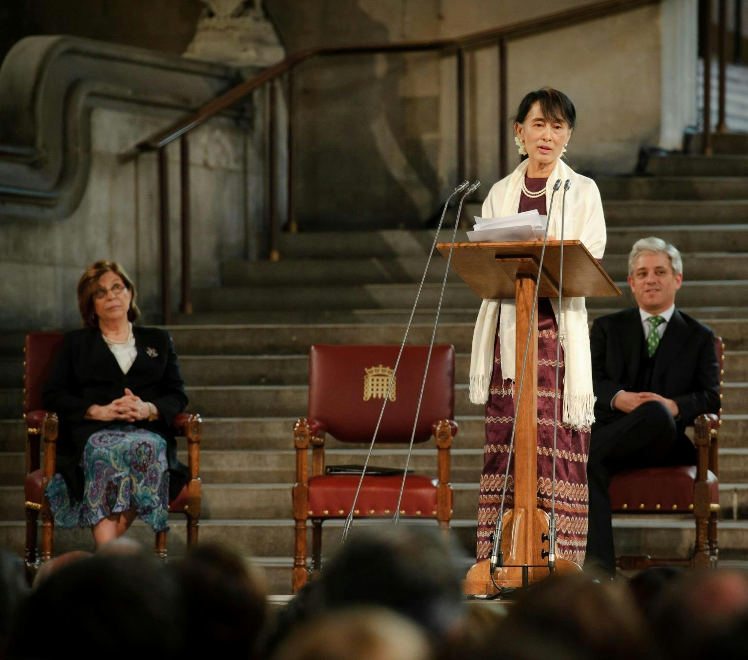 Aung San Suu Kyi is weraing a dark purple dress and a white shall. She is standing at a wooden lecturn and is mid speech. Behind he is a woman and a man stting in chairs watching her speak.