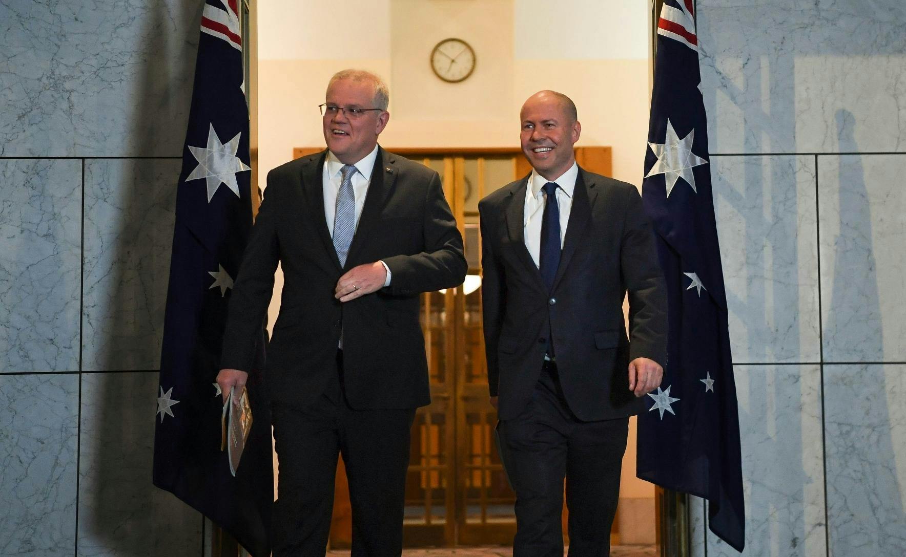 Scott Morrison and Josh Frydenburg wearing black suits. The are smiling and walking out of a room. Two Australian Flags are hung behind them.