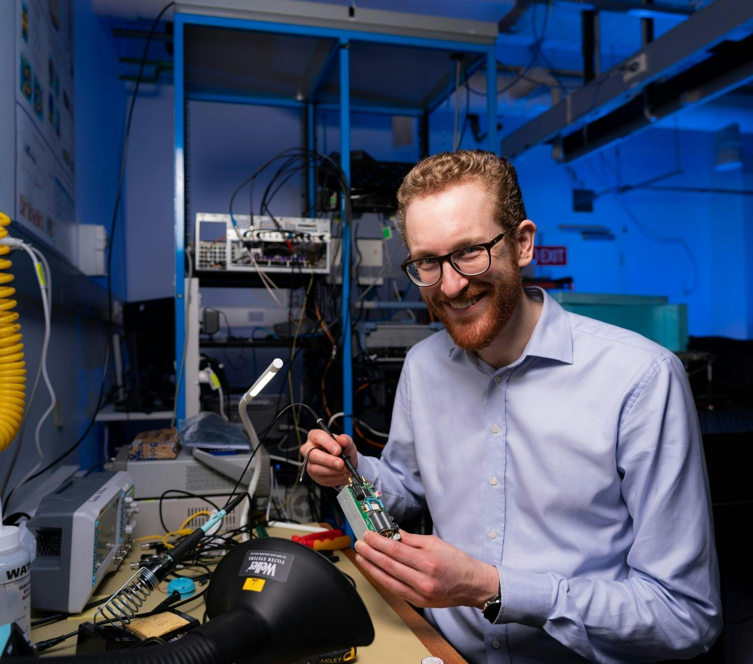 Andrew Horsley is wearing a blue collared shirt and black glasses. he is smiling at the camera and holding a small box covered in buttons and wires. He is in a room filled with various technology items and blue light.