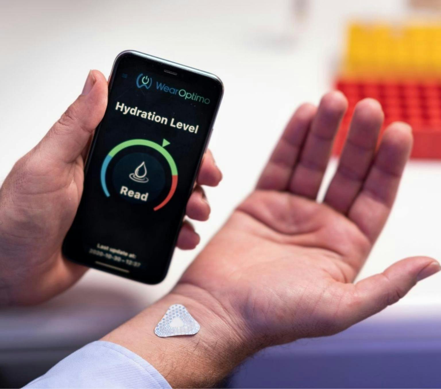 close-up of a persons hand. they are holding a smart phone which displays a dehydration level barometer in their left hand. Their right hand is facing palm up and a small triangle sticker is placed on their right wrist. The sticker is the Wearoptimo micro sensor.