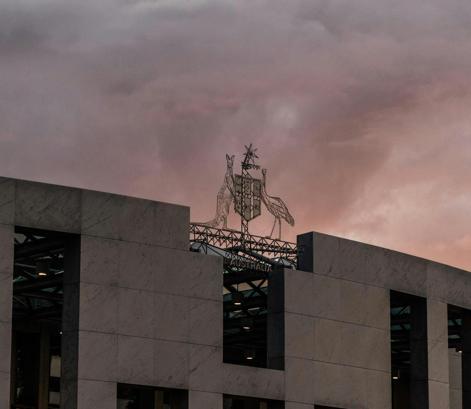 The Australian coat of Arms featuring a shield, an emu and kangaroo above parliament hosue are pictured above a pink sky at dusk.