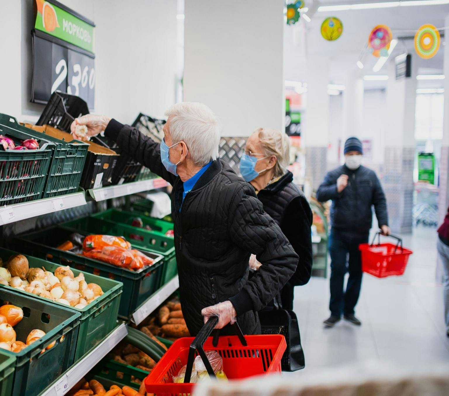Man and woman with white hair, black hair and blue masks. They are facing a wall of fruit and vegetables in a supermarket. The man is reaching for a bulb of garlic.