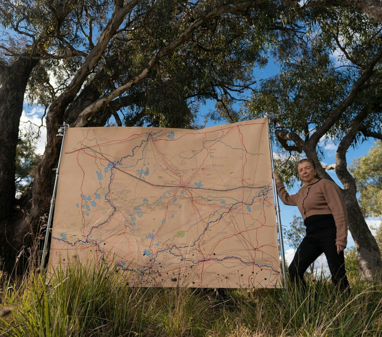 Ann is wearing a brown jumper and black pants. She is standing next to a large light brown map which is taller than her. She is outside, standing in grass with tall trees behind her