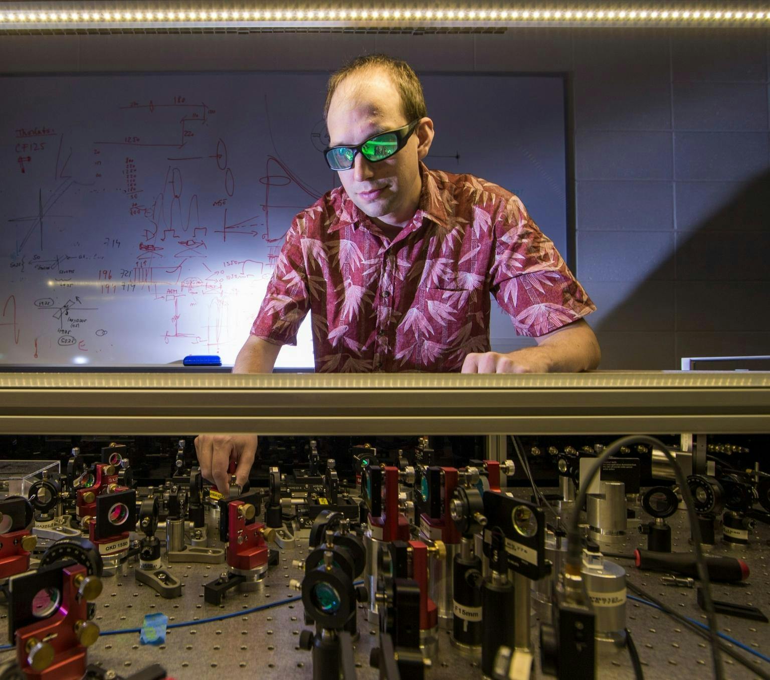 Bennet is wearing sunglasses with reflective green lenses and a pink floral collared shirt. He is leaning over a desk covered in small technology objects. A whiteboard with scientific diagrams is behind him.