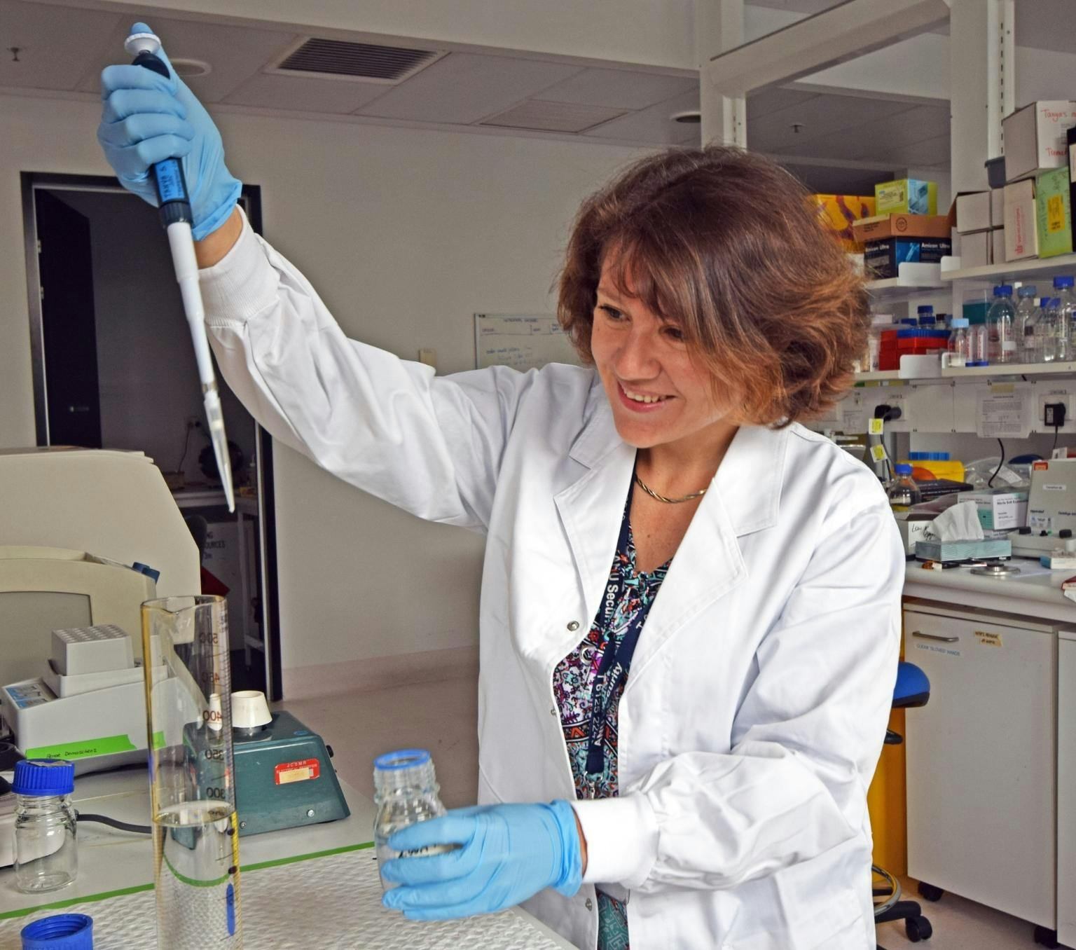 Tanya is holding a long pipette tube and dropping liquid into a measuring cylinder. She is wearing a white lab coat and standing in a laboratory.