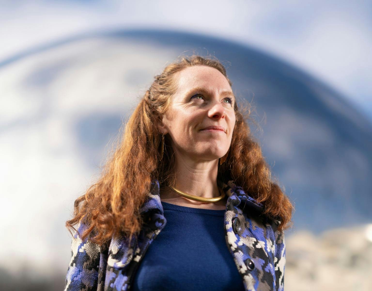 Cassandra Steer, a woman with long red curly hair, looks up and away from the camera while standing in front of a large dome-shaped building.