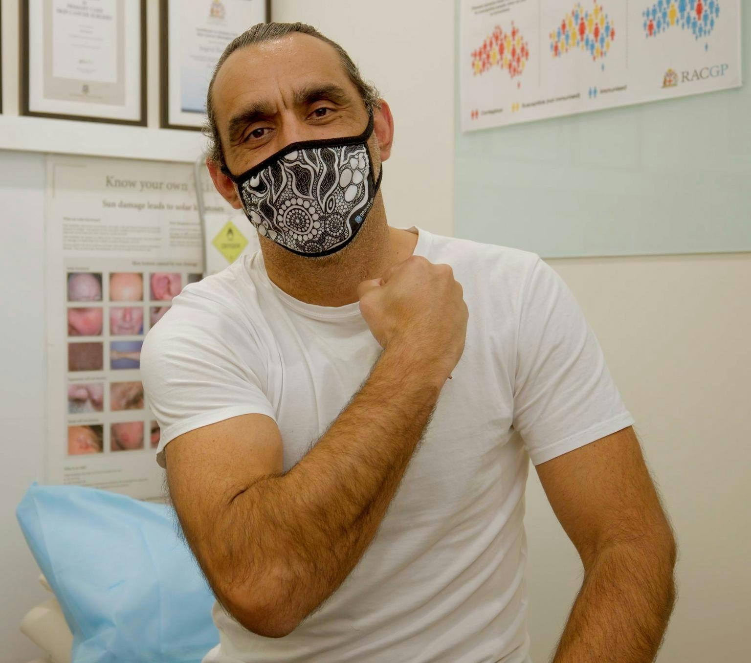 Adam Goodes is wearing a white Tshirt. He is wearing a mask with a black and white Indigenous artwork. His right arm is held across his chest. He is sitting in a room surrounded by medical posters.