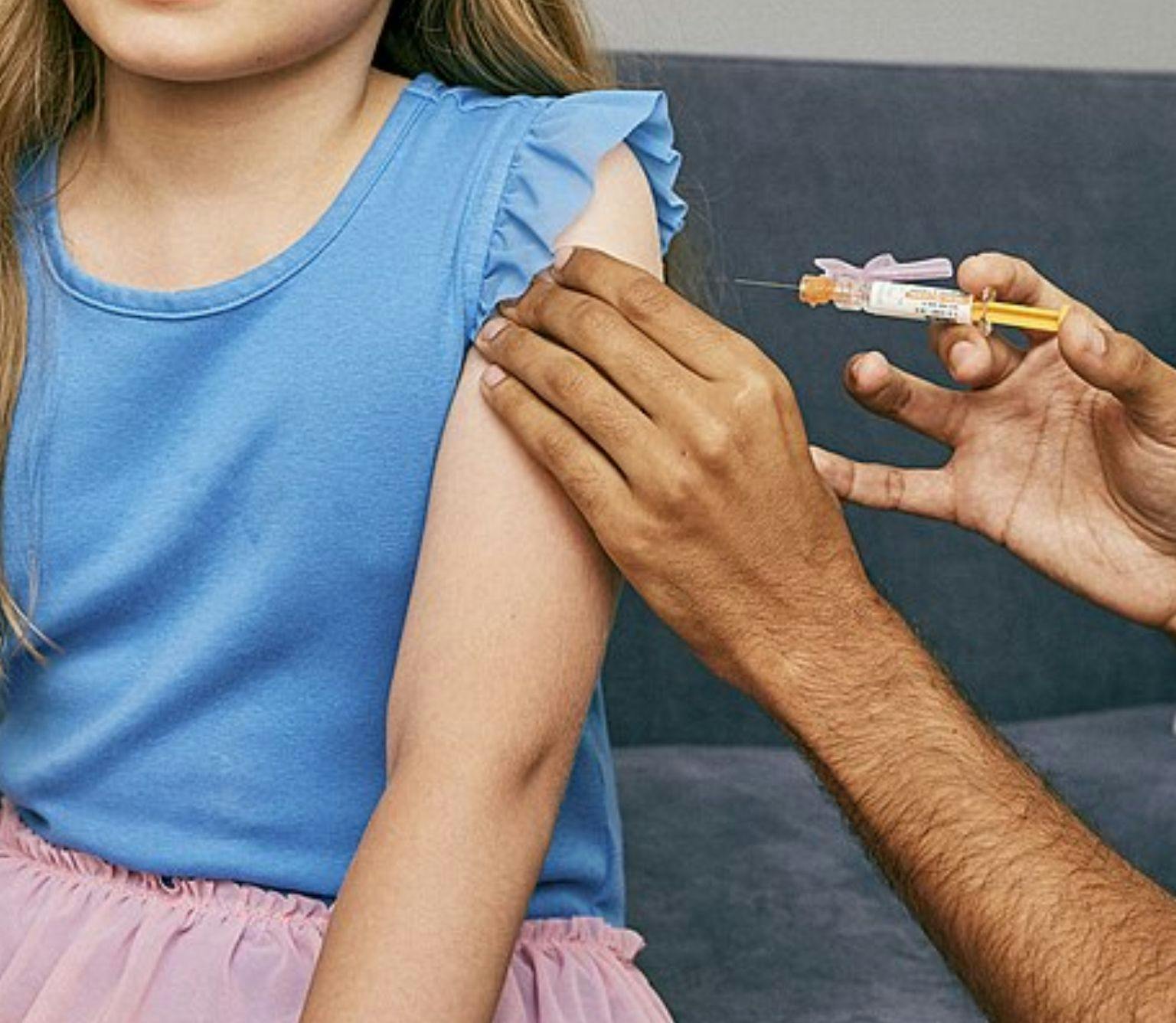 The torso and arm of a child in a blue shirt is visible. An adult is holding her arm with one hand and holding a vaccine syringe in the other hand.