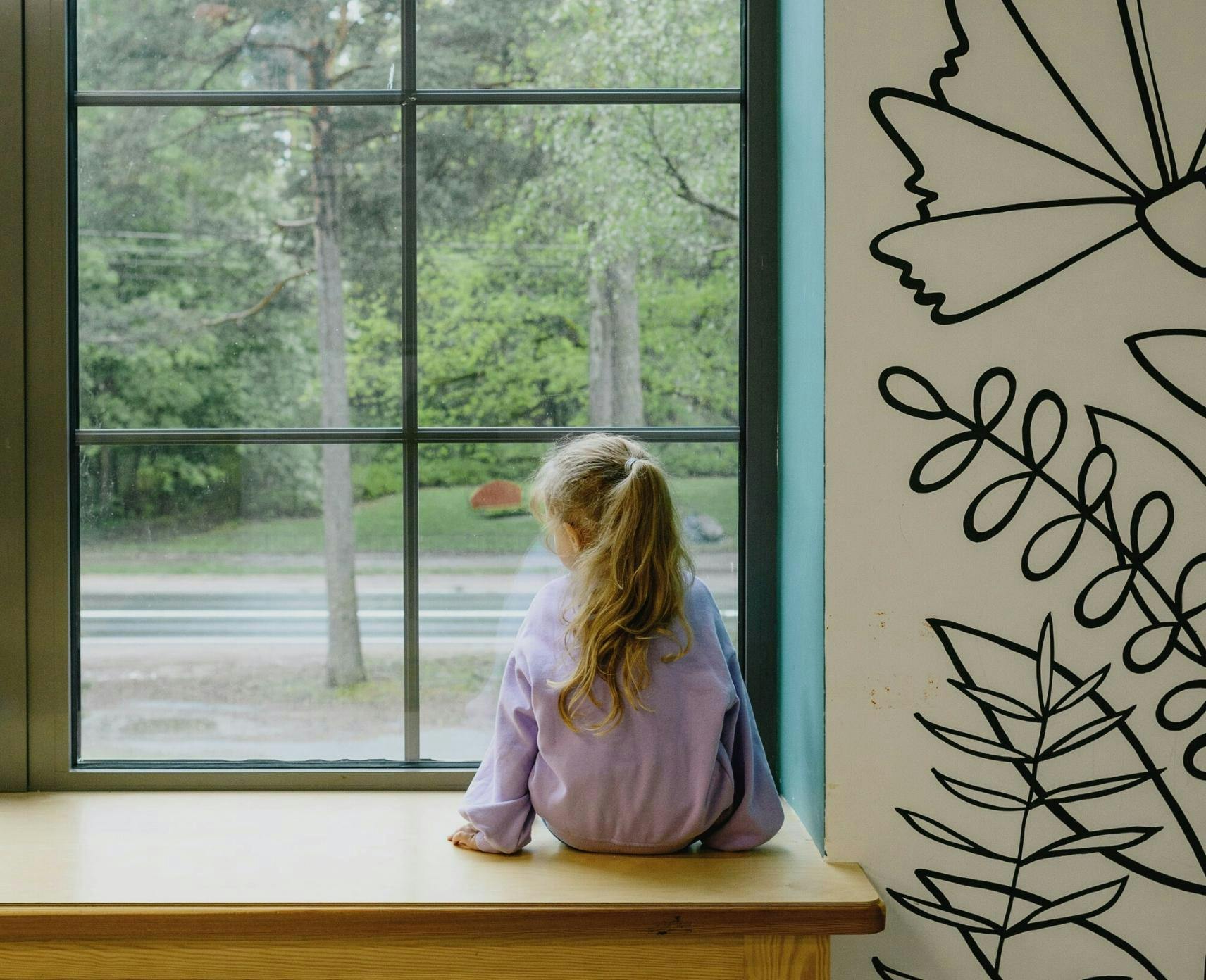 child in lilac jumper is sitting down with her back to the camera. She is looking out a window which shows trees and a street.