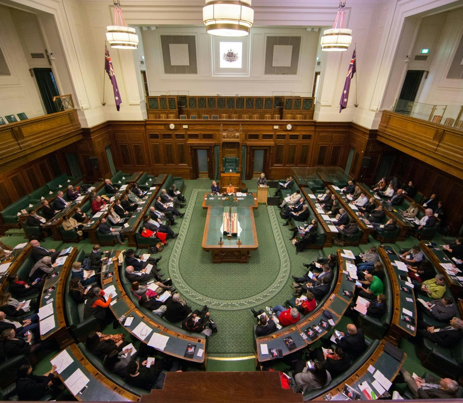 Looking down on the House of Representatives chamber in Old Parliament House. There is a green carpet on the floor, with wooden desks and green leather chairs in a 'u' shape around the centre desk. The seats are filled with people sitting in them.