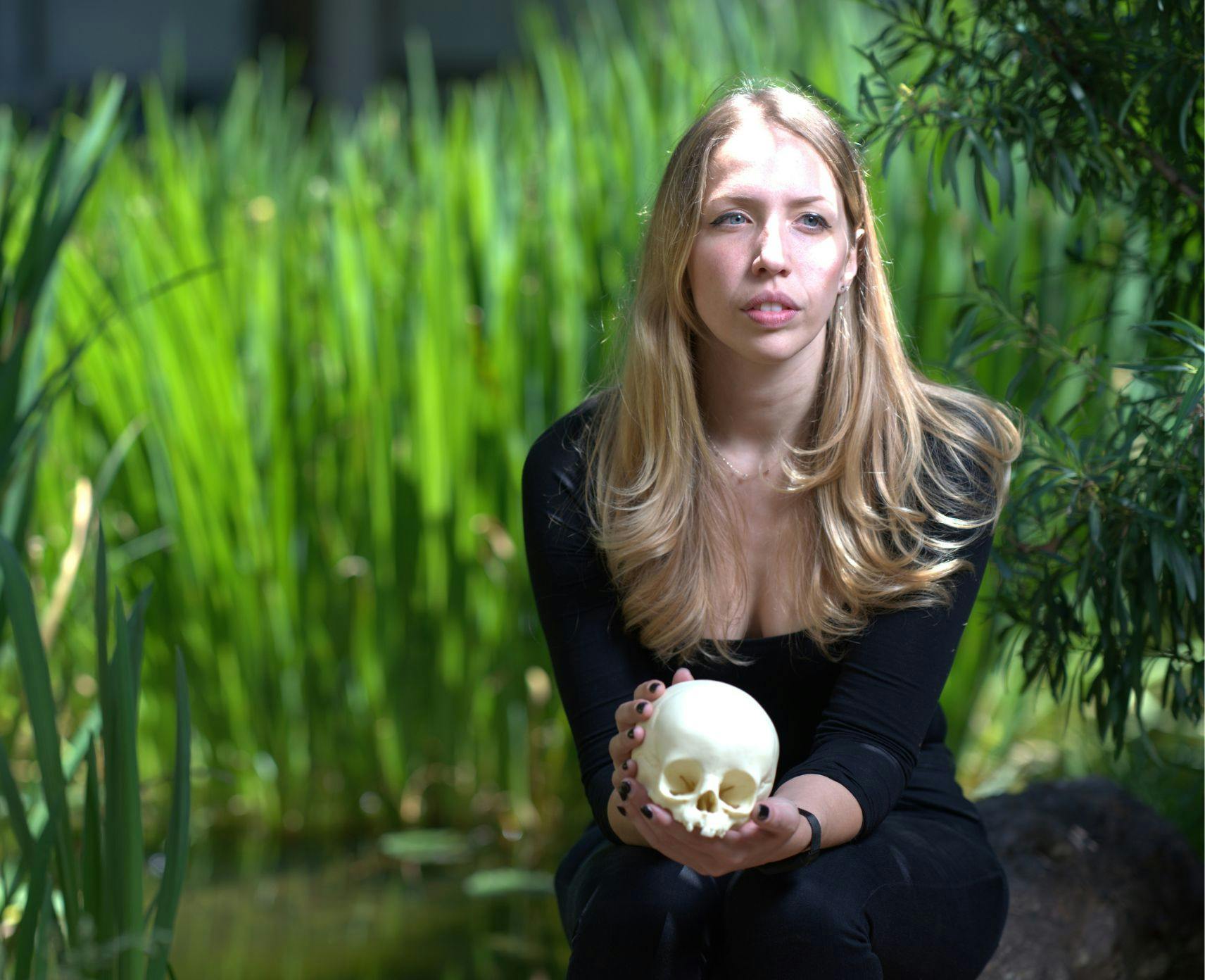 Dr Clare McFadden holding a human skull. She is crouched between tall grass.
