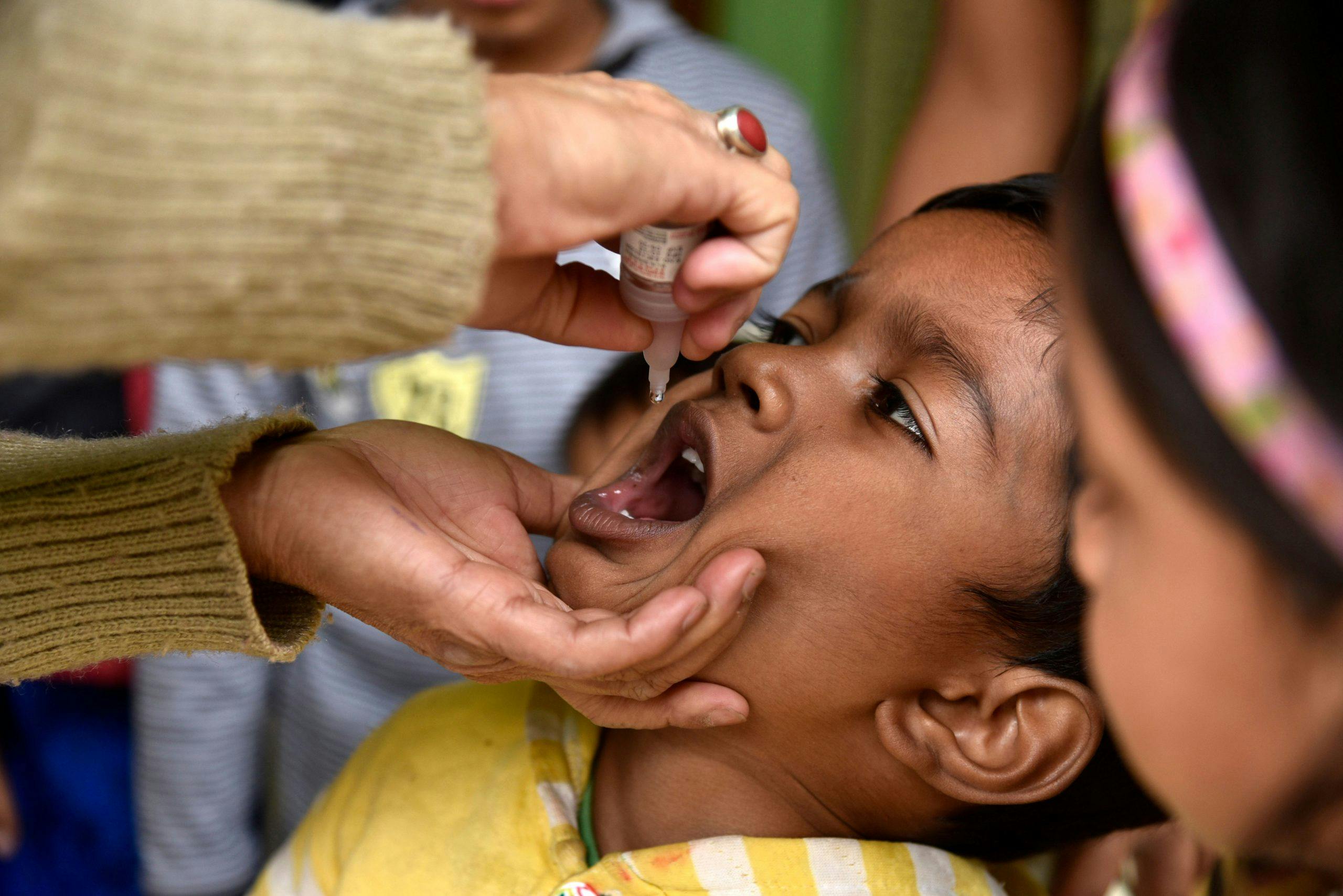 An Indian child tilts their head up as an adult's hand squeezes a liquid vaccine into the child's mouth.