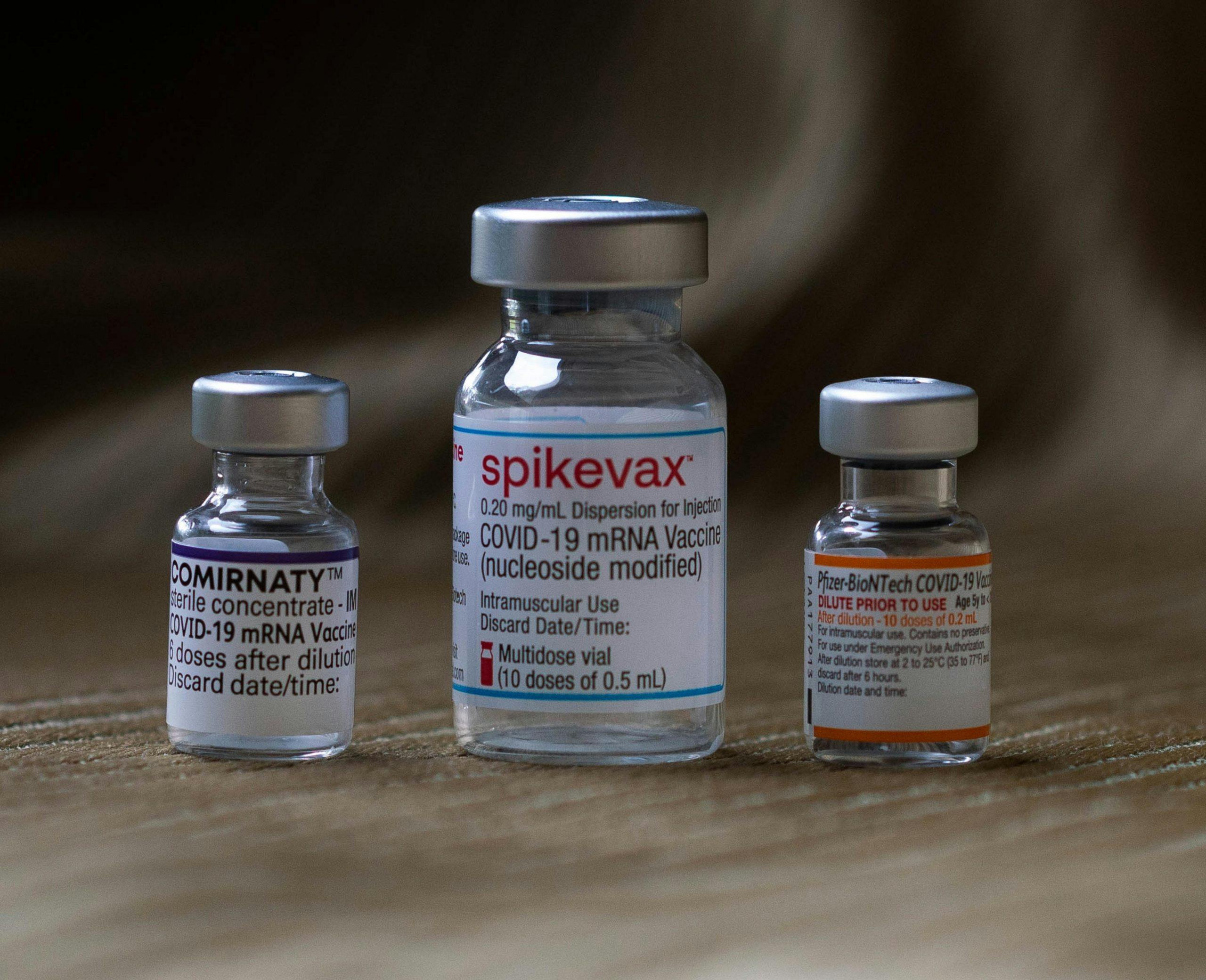 close up shot of three covid vaccine vials. They are labelled: Cominaty, Spikevax and Pfizer-BioNTech (left to right)
