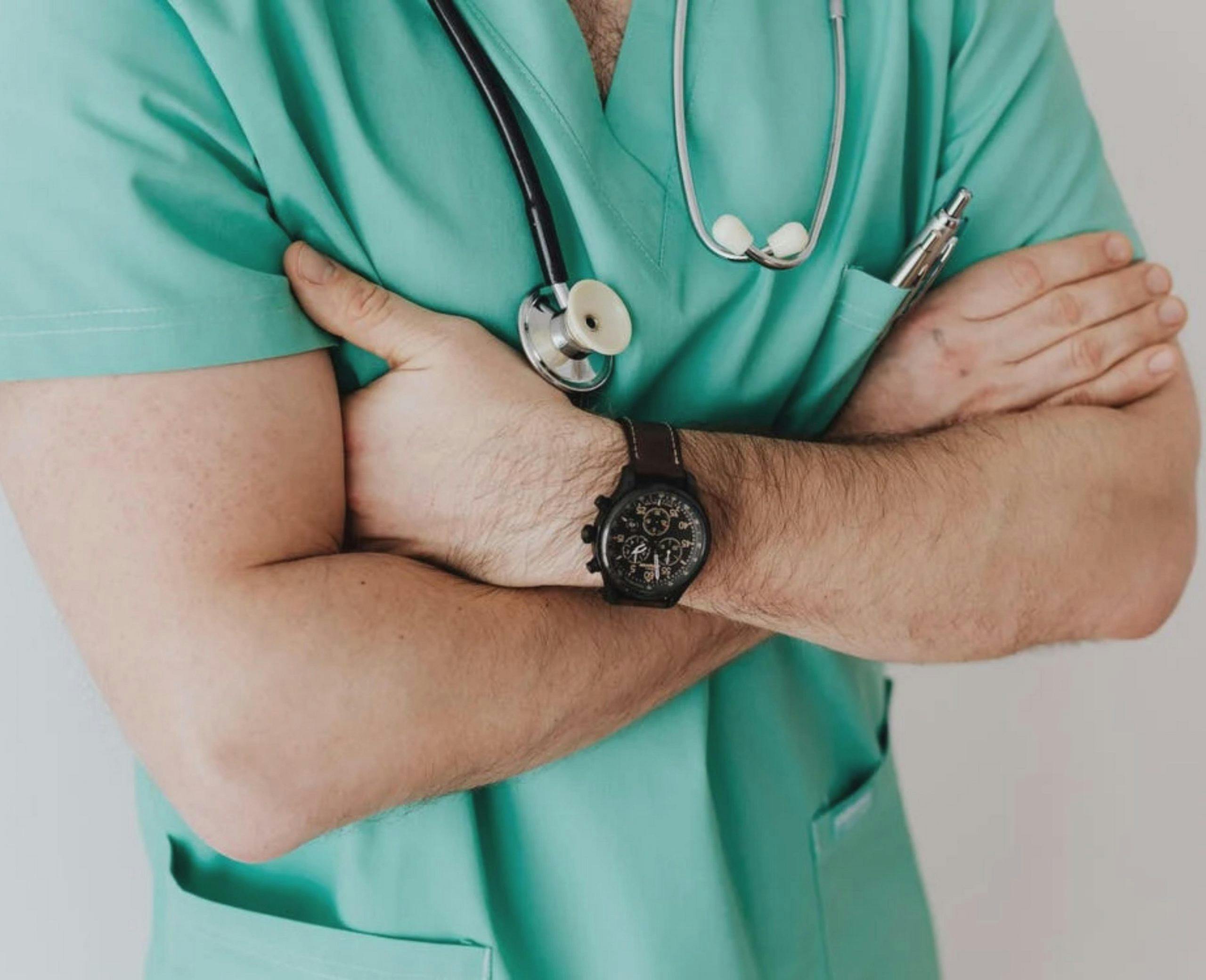 Close-up photo of a man's torso with his arms folded. He is wearing green hospital scrubs, and has a stethoscope around his neck.