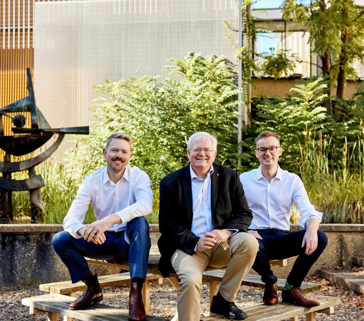 THree men are in business casual clothing and sitting on a picnic table bench smiling at the camera