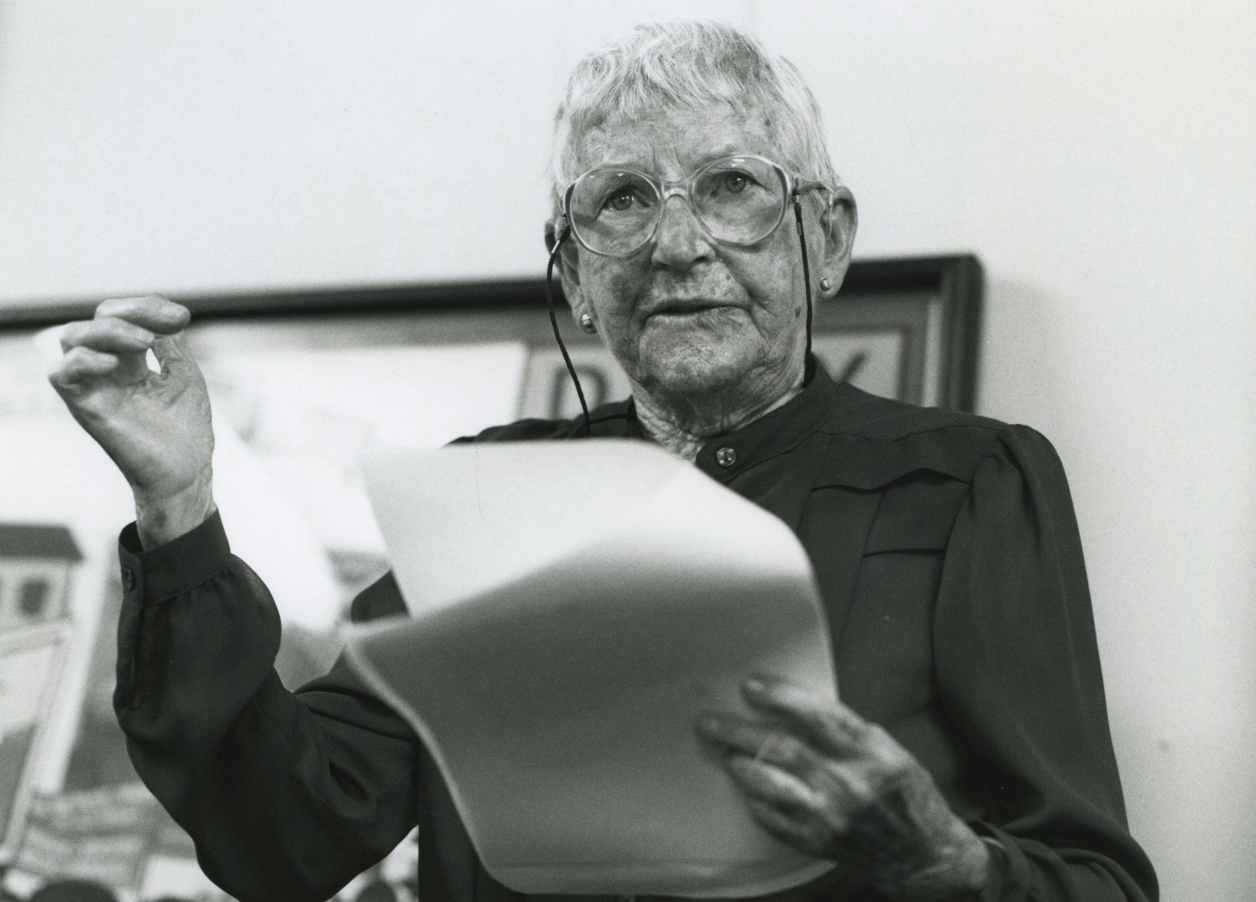 A black and white photo of Edna Ryan, a woman with short white hair and wearing glasses, and holding a piece of paper from which she appears to be reading from as she speaks.