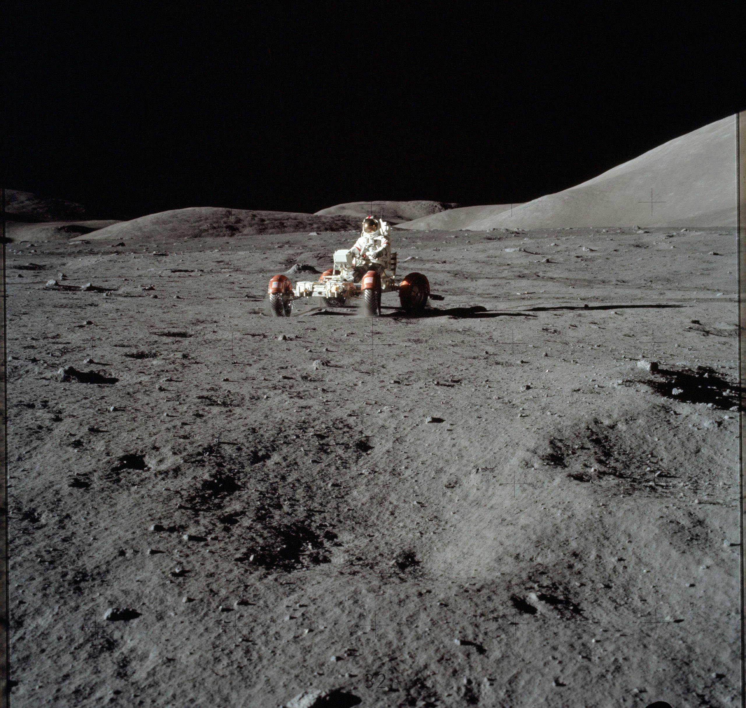 An astronaut in a space suit sits on top of a red four-wheeled buggy on a grey rocky and sandy landscape.