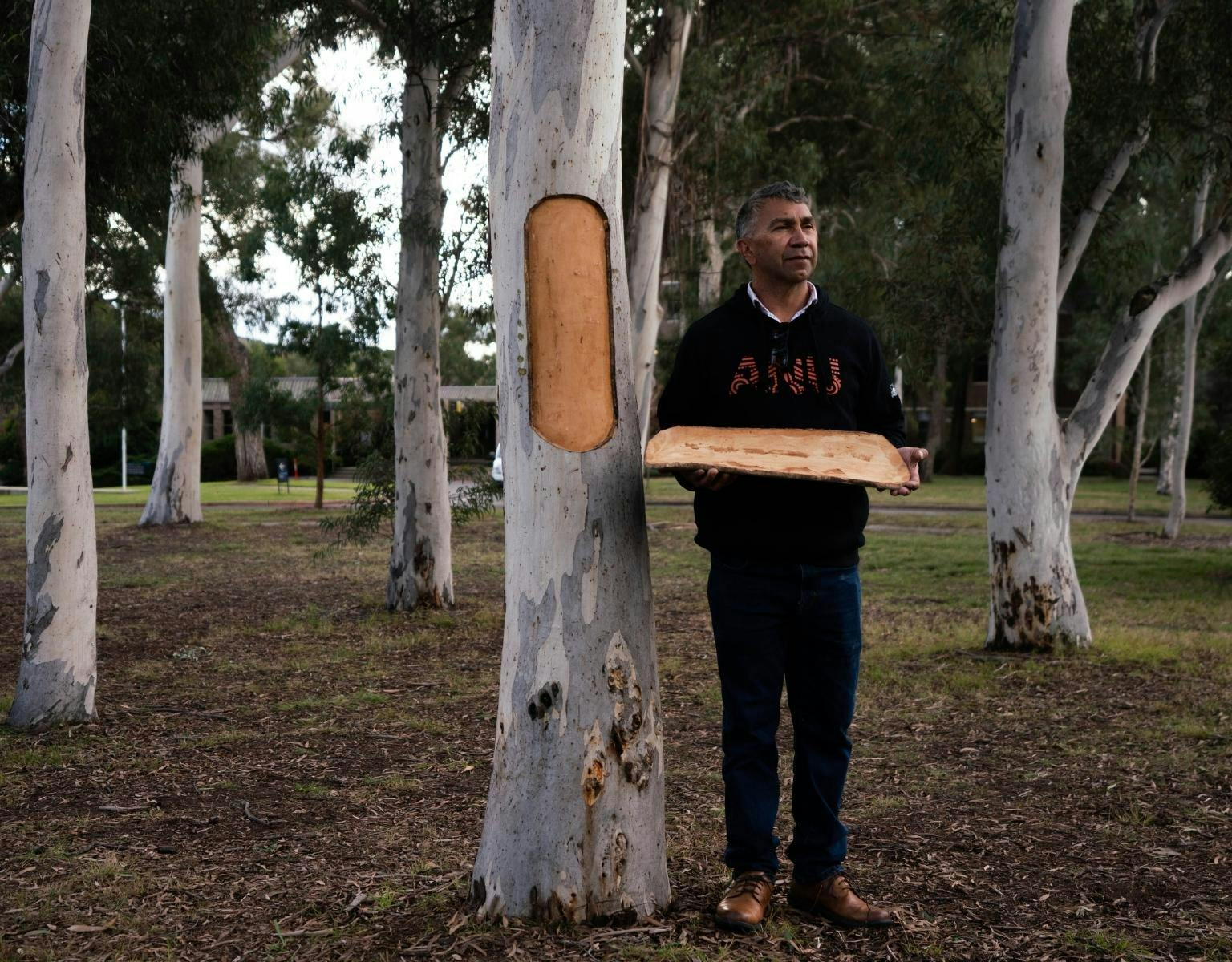 Paul Girrawah House, wearing a black jumper with the letters A N U on the front, stands next to a tree holding a piece of bark he has carved out of the tree.