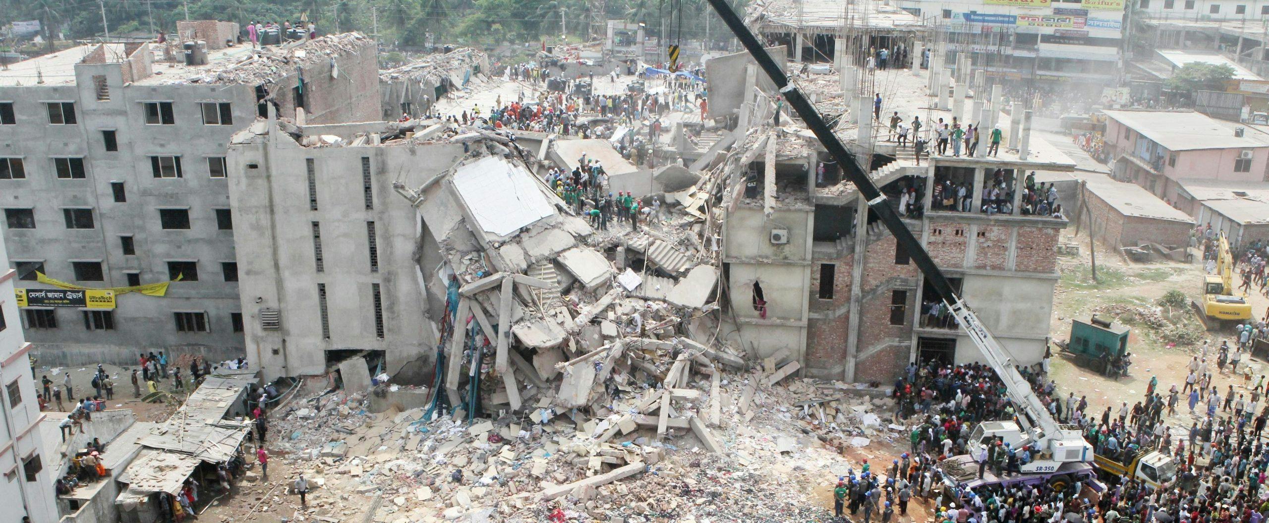 Rescuers climb over debris from a large collapsed building. A large crowd of people gather round a crane, which is being used to remove pieces of debris.