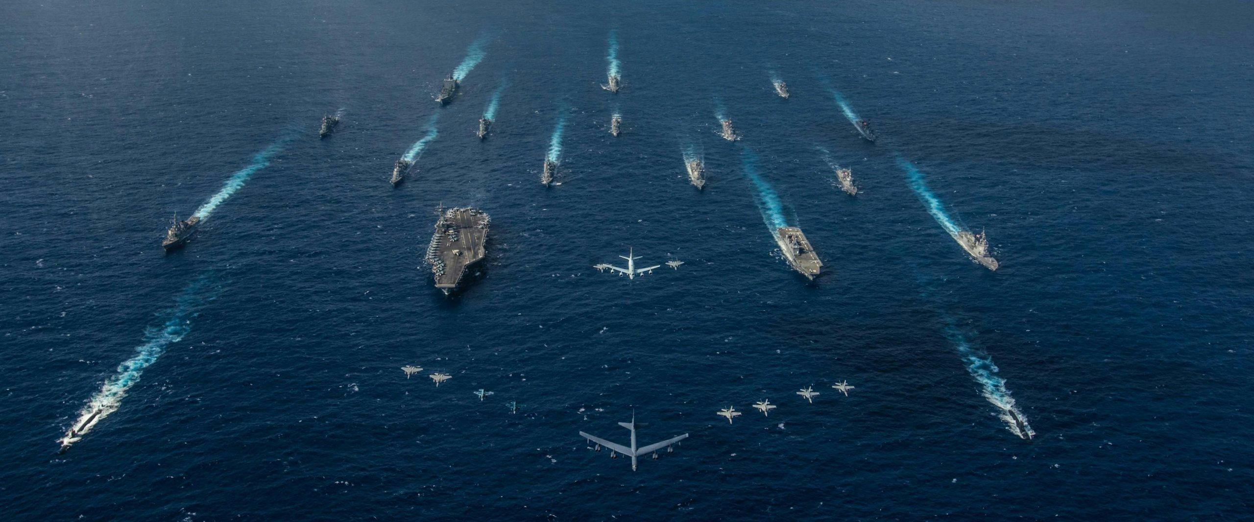 Ariel view of navy ships in the ocean and aircraft flying above them.