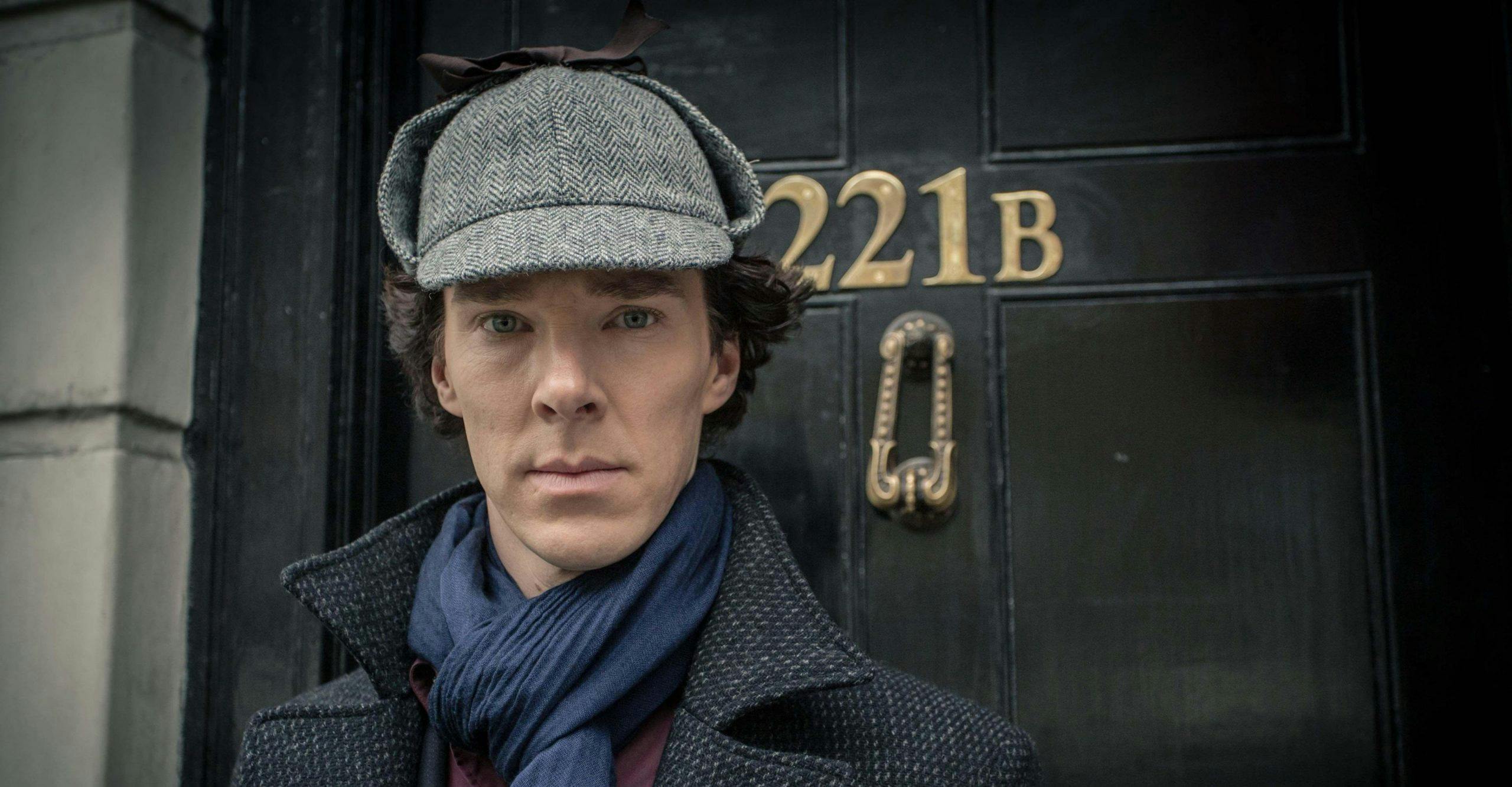 Benedict Cumberbatch as Sherlock Holmes wears a grey deerstalker cap, navy scarf and dark grey jacket and stands in front of a black door that has the address 221B written in gold.