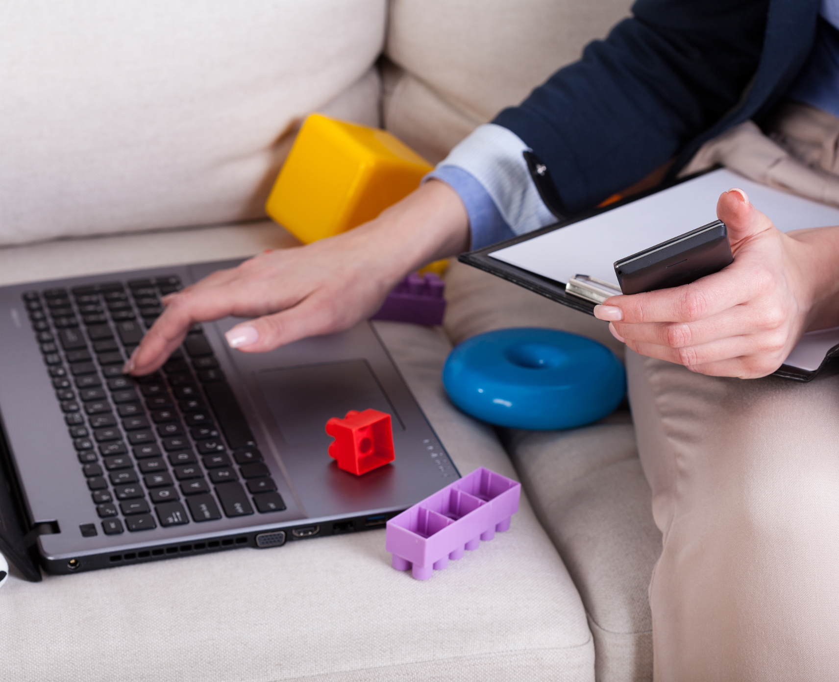 A woman's hands are shown holding a smartphone and touching a laptop. She is seated on a couch next to children's toys.