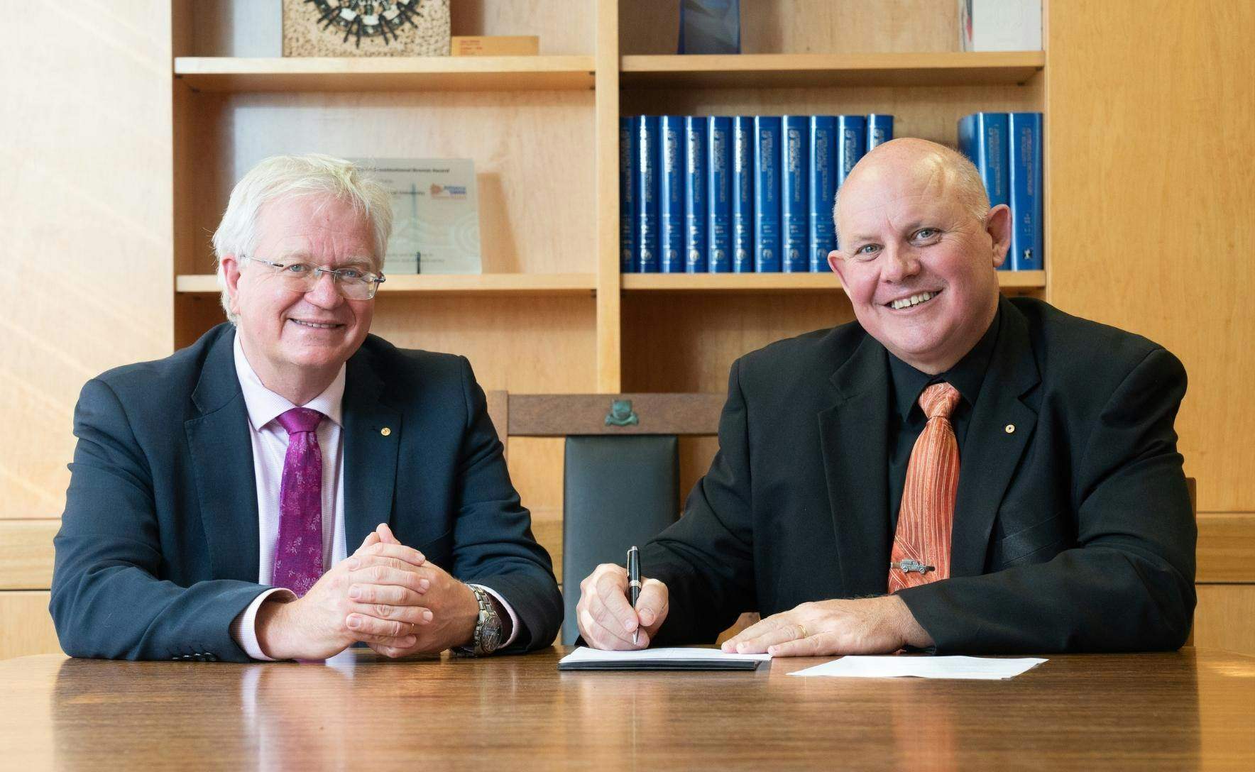 Professor Brian Schmidt and Professor Scott Bowman sit at a large table. Professor Bowman holds a pen in his right hand over a piece of white paper on the table.