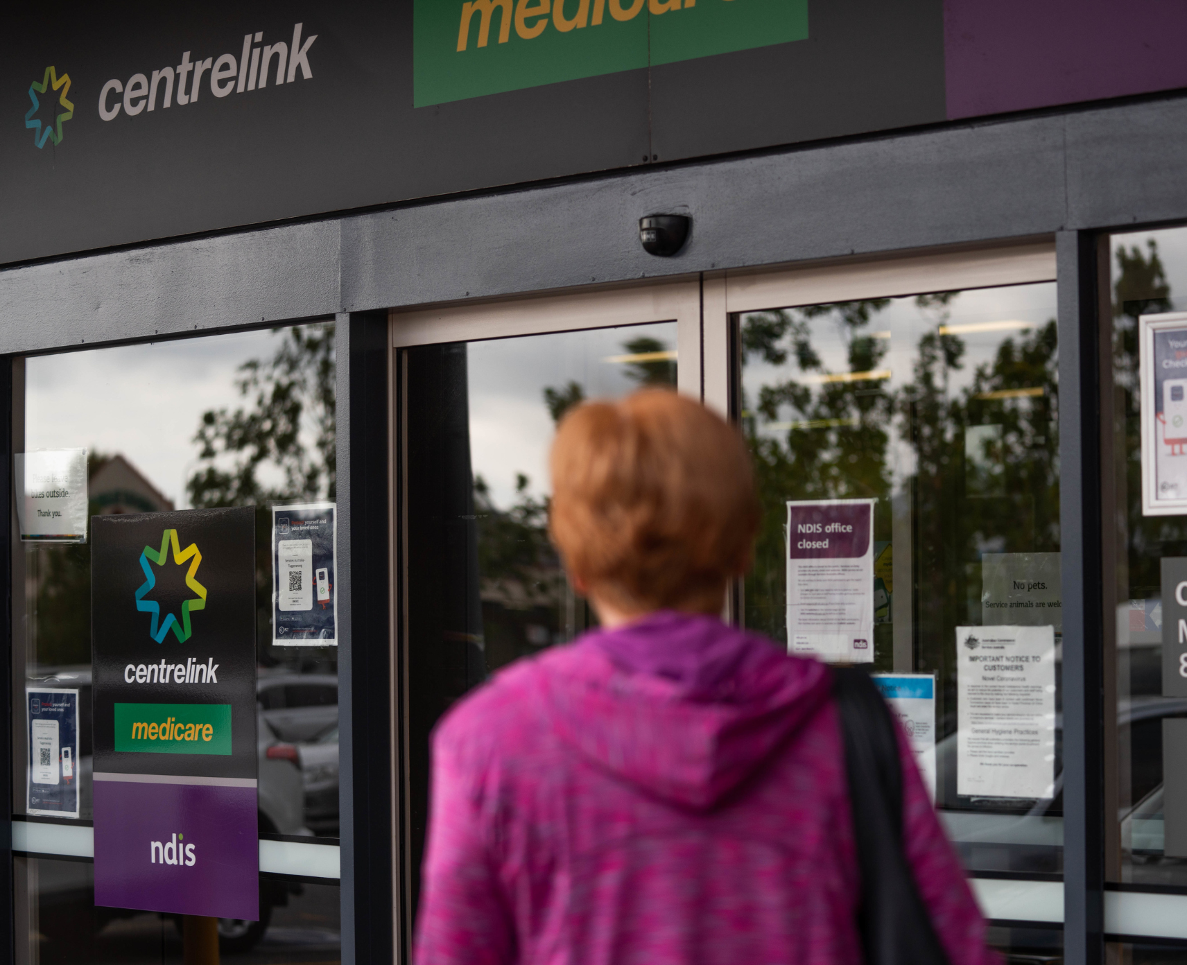 A woman with short red hair is pictured from behind walking towards the entrance of a Centrelink office.