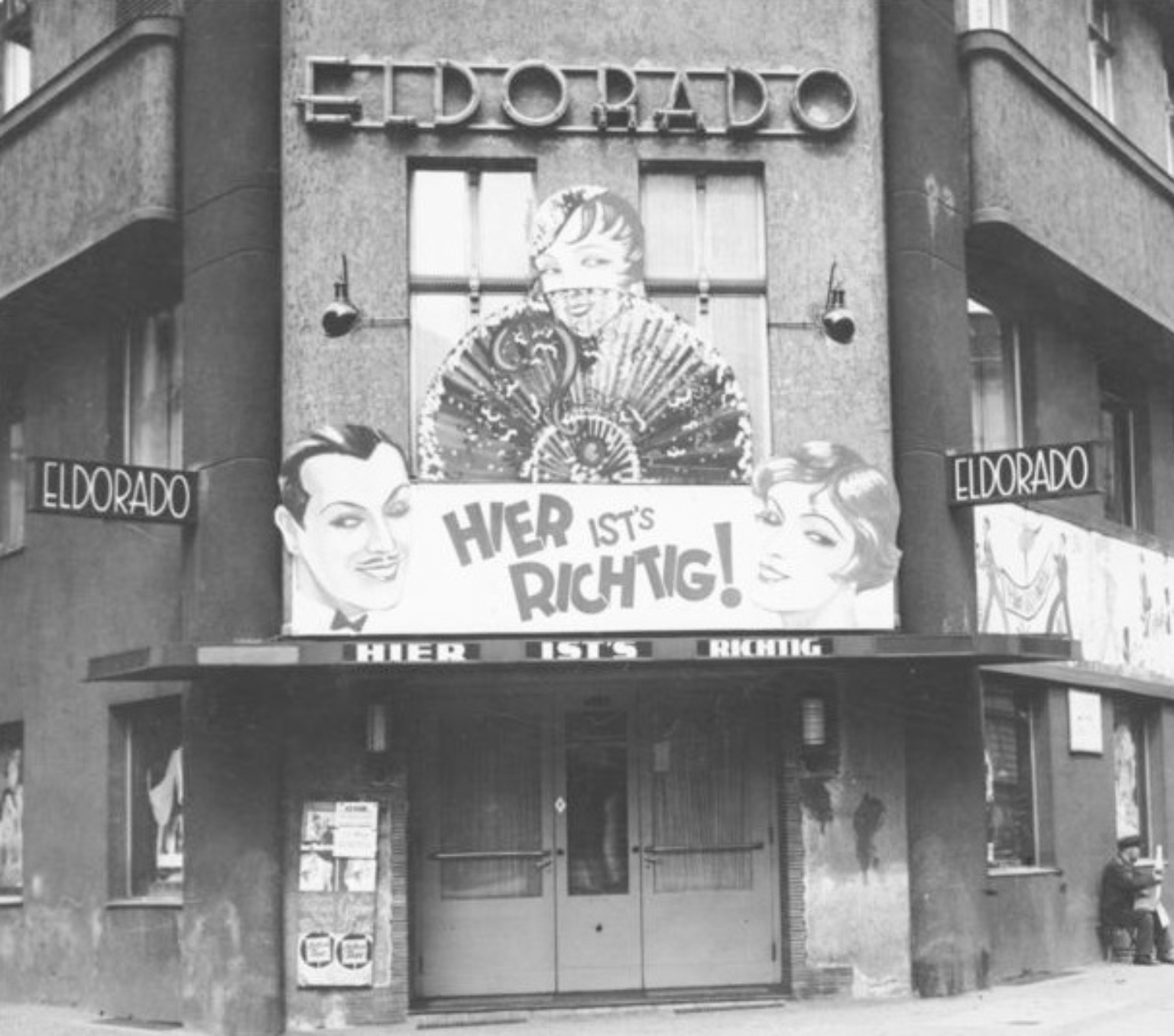 A 1930's nightclub with the name Elorado signposed high on the facade. A banner with the faces of a man and a woman has the phrase "Hier Ist's Richtig" above the door.