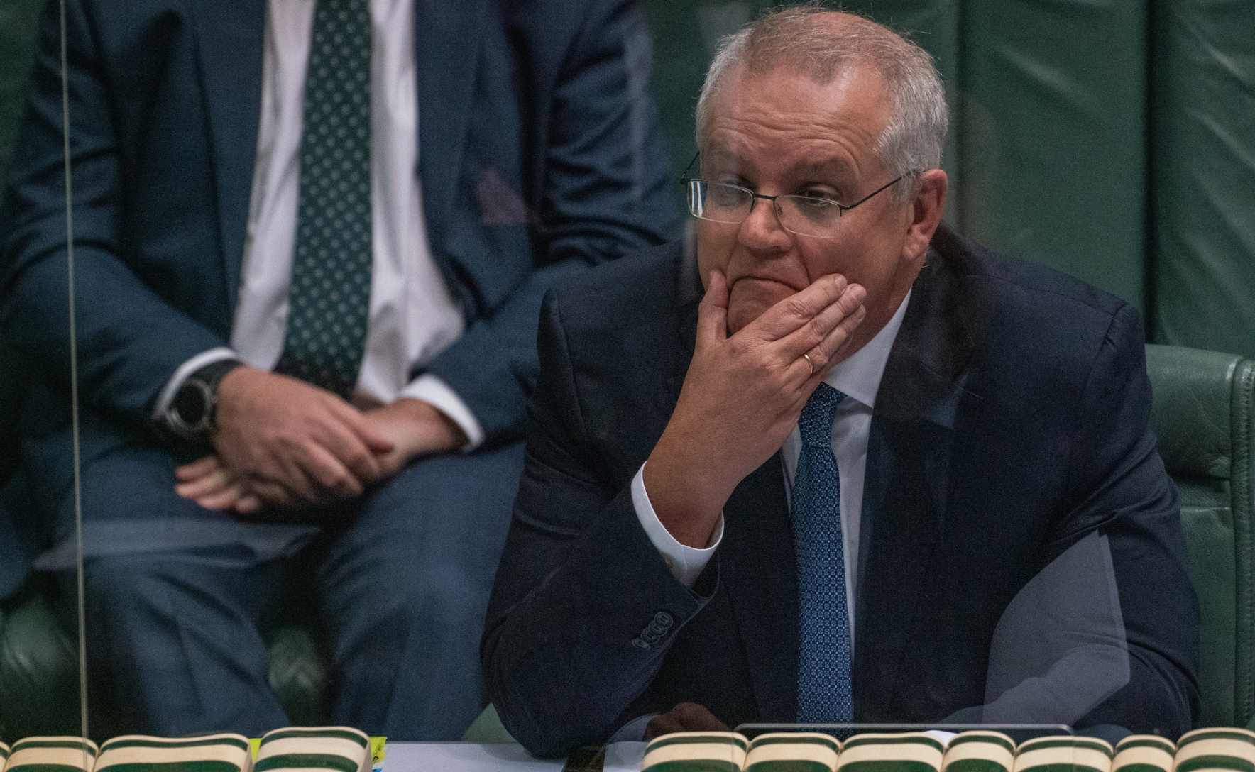 Prime Minister Scott Morrison sitting in parliament behind a sheet of plexiglas. He is grimacing and has a hand on his chin.
