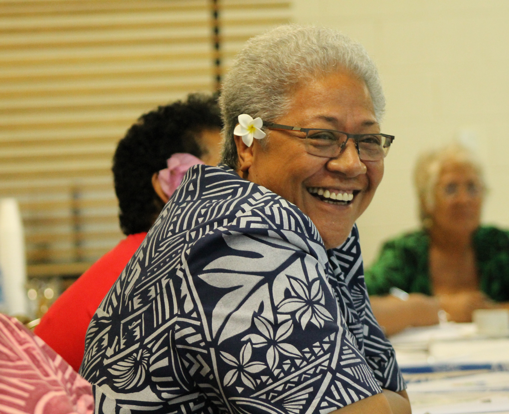 Fiame Naomi Mata’afa, a woman with short, grey hair, is seated at a table with a small white flower tucked behind her ear.