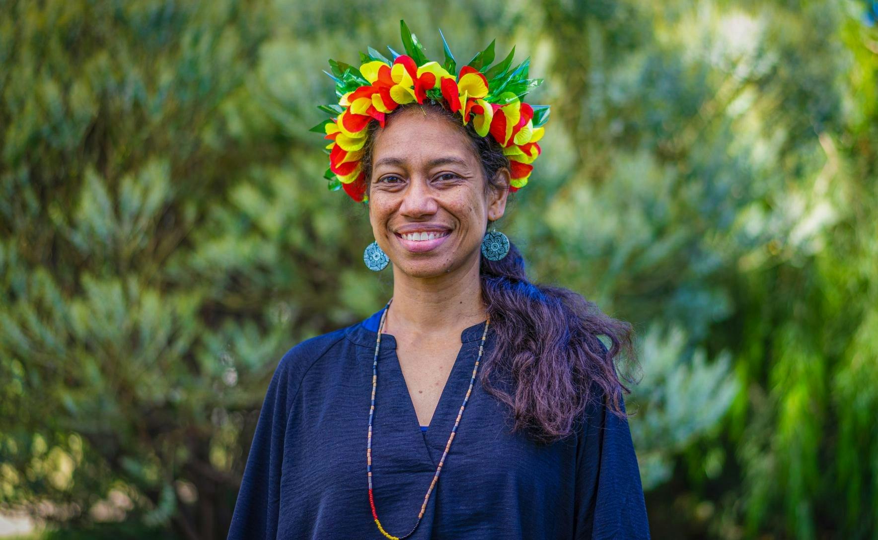 Katerina Teaiwa wears a yellow, red and green head piece, a beaded necklace and bright blue patterned earrings. She stands in front of bright green plants.