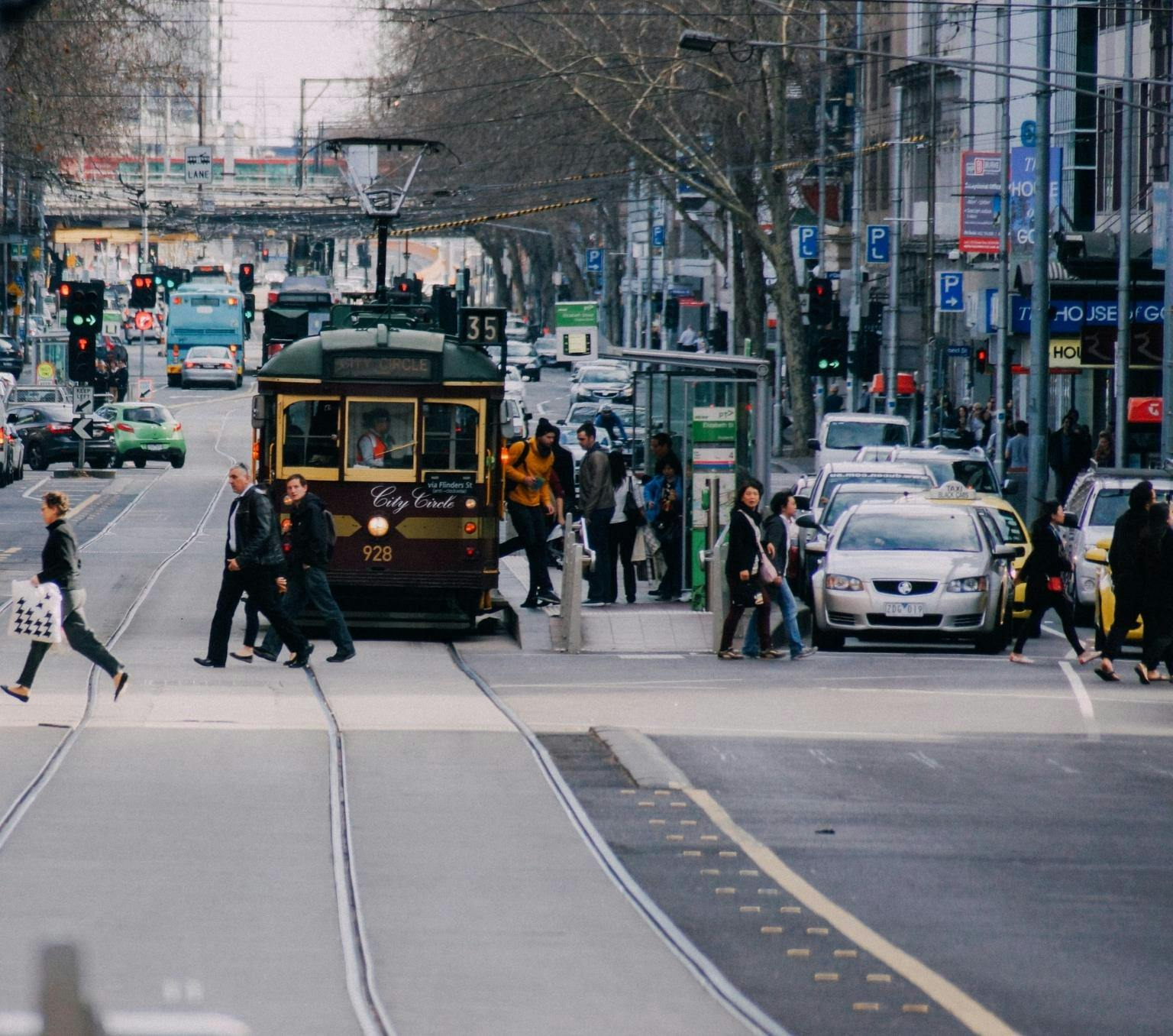 Streetscape of a melbourne street. There is a yellow and green tram, cars on the street and people crossing the road.