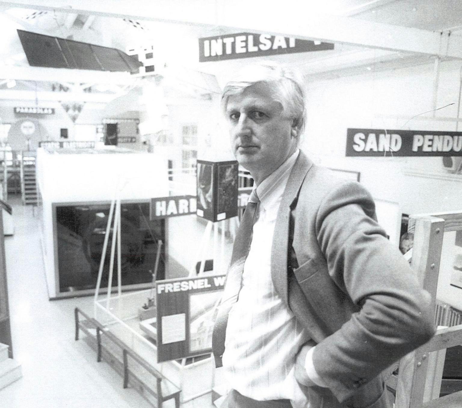 Professor Michael Gore AO in a hardware store. He is wearing a shirt, tie and blazer. There is a sign behind him that reads: 'sand pendulum'. The photo is black and white.