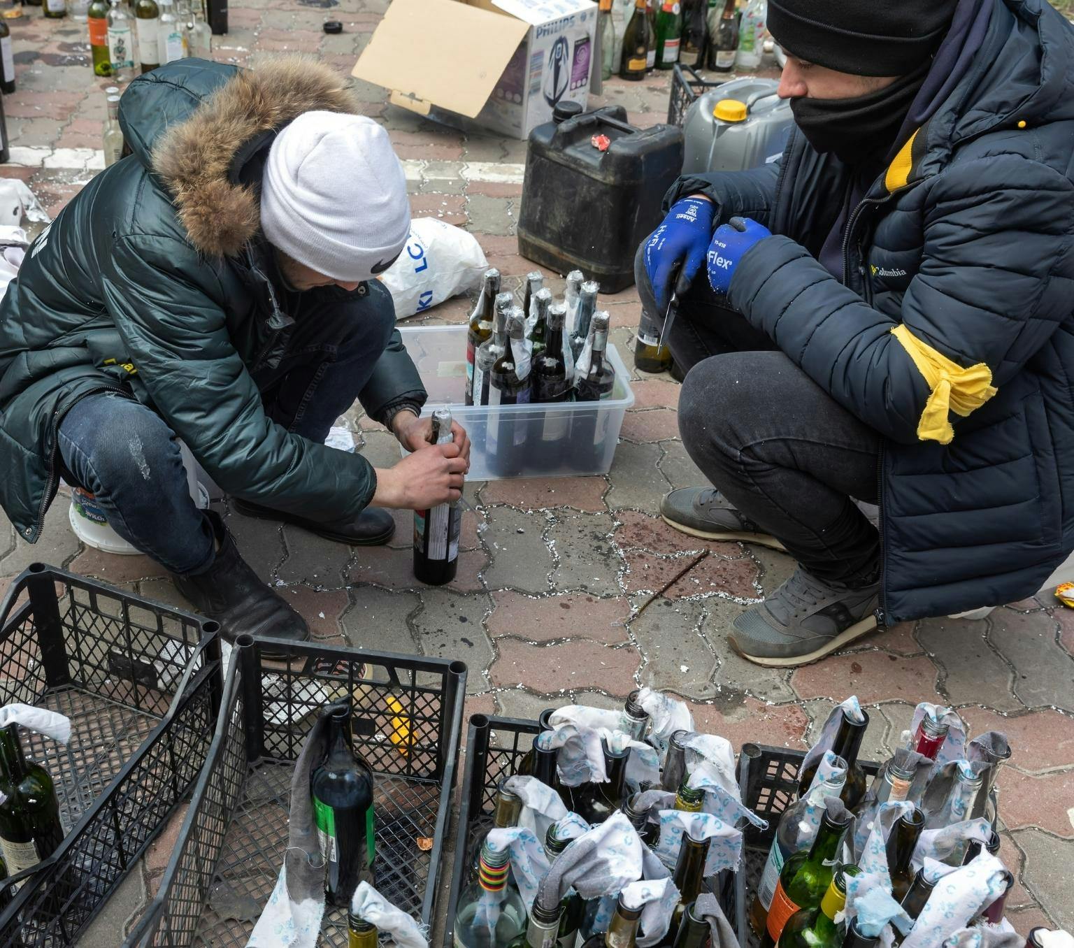 Two people wearing beanies and dark puffer jackets are crouched on the ground. One is holding a tall brown bottle. On the ground in front of them are small black crates filled with bottles with white cloths coming out of the top. In the background are plastic fuel storage containers and empty wine bottles.