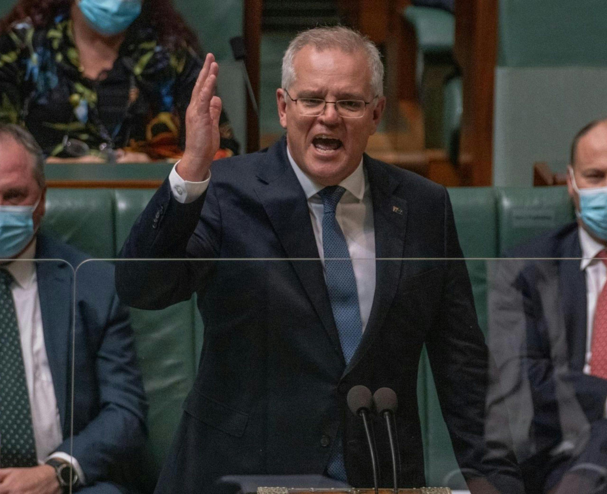 Scott Morrison standing in the lower house of Parliament with Barnaby Joyce and Joshua Frydenburg sitting behind him in masks and suits. Morrison has his right hand raised and is mid-speech