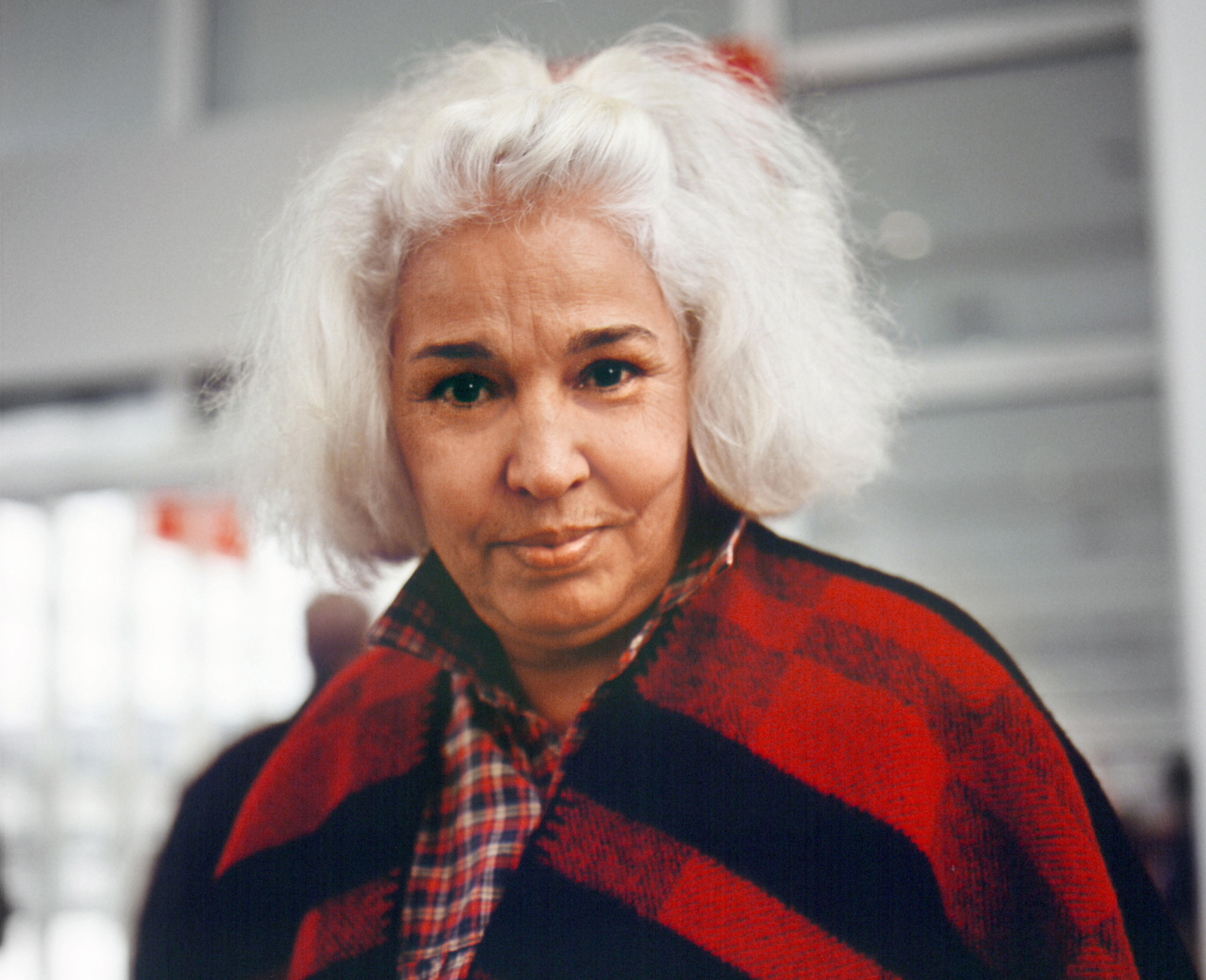 Nawal El Saadawi, a woman with a bob of white hair, looks at the camera wearing a black and red check coat.