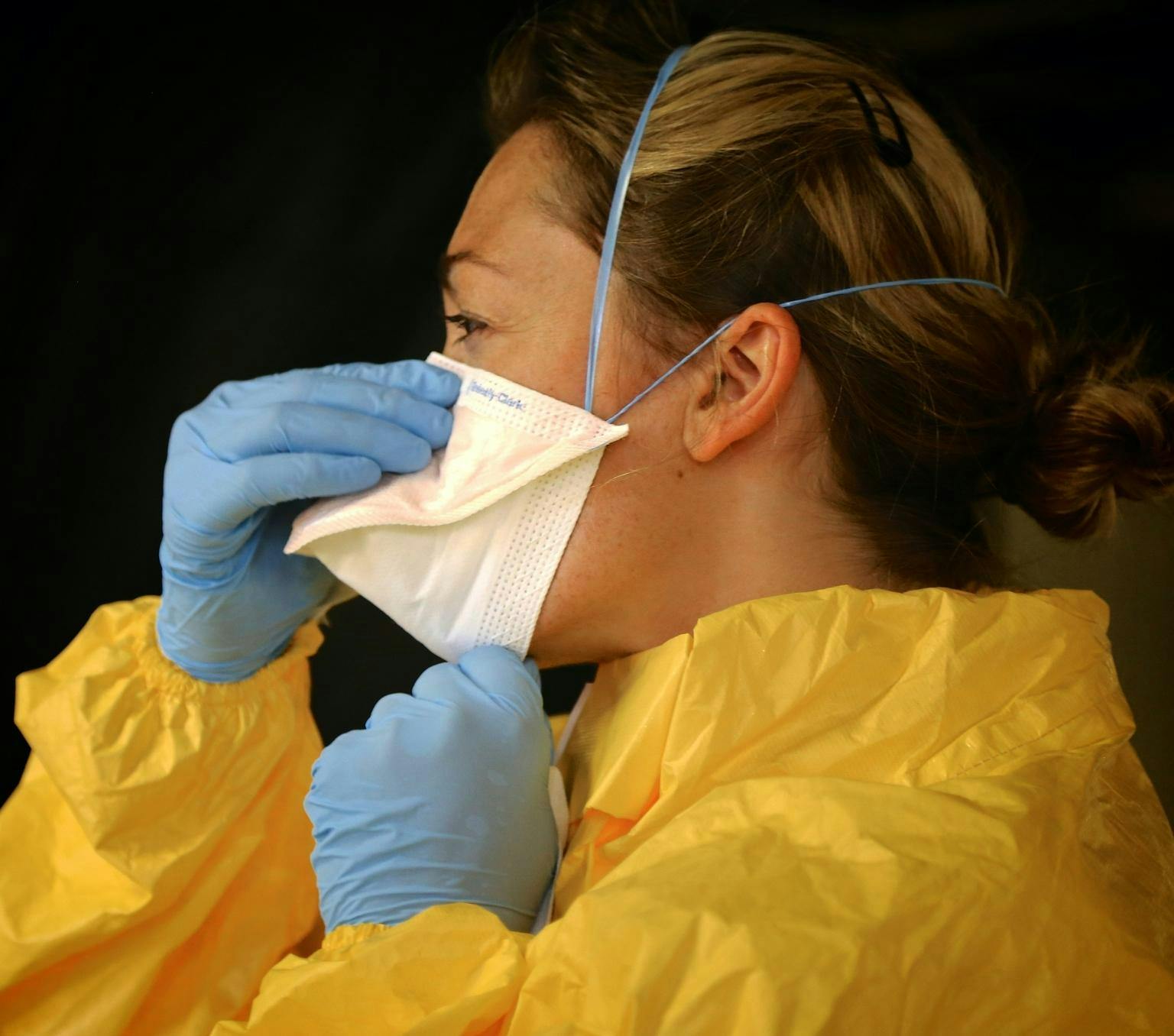 profile of a woman wearing a yellow mazmat suit, blue plastic gloves and a white face mask. She is adjusting the facemask with her hands.