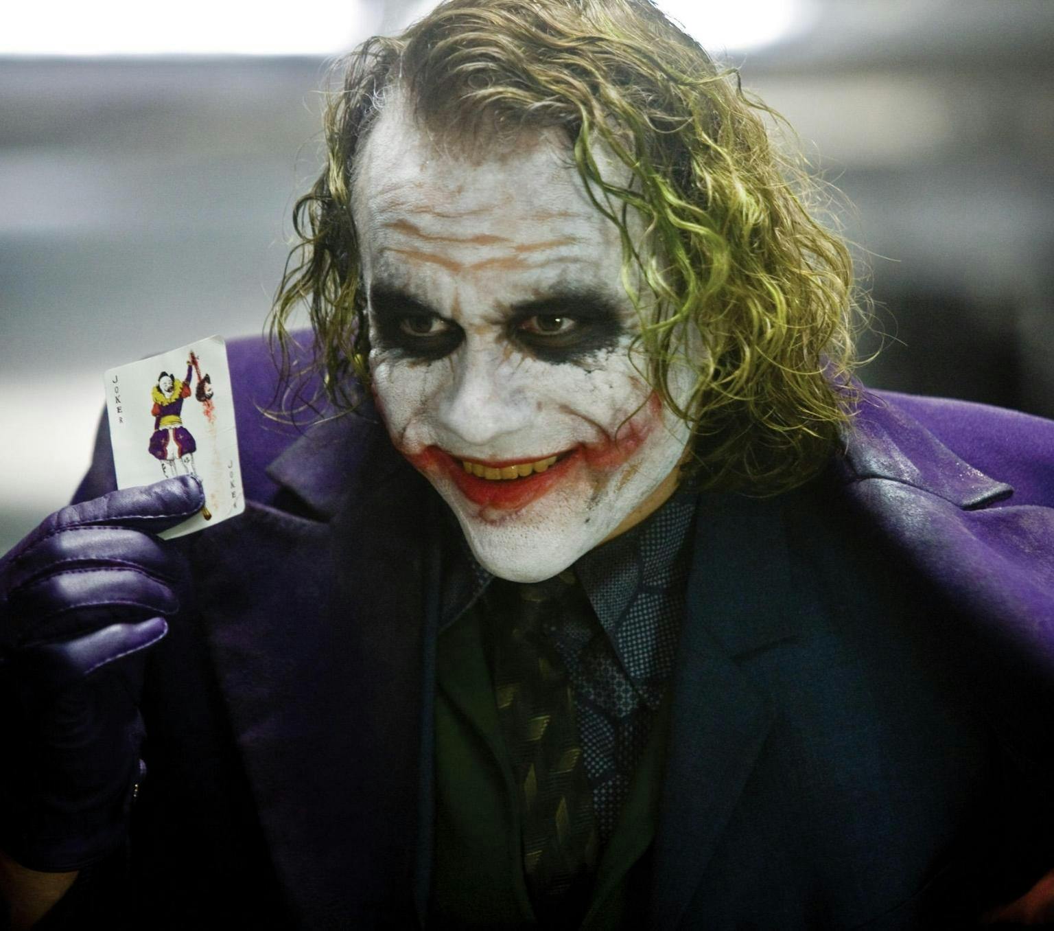 Heath Ledger as the Joker, with yellow hair, white make-up on his face, and red lipstick smudged on this lips and across this cheeks. He wears a purple jacket and gloves and holds up a playing card featuring the joker.