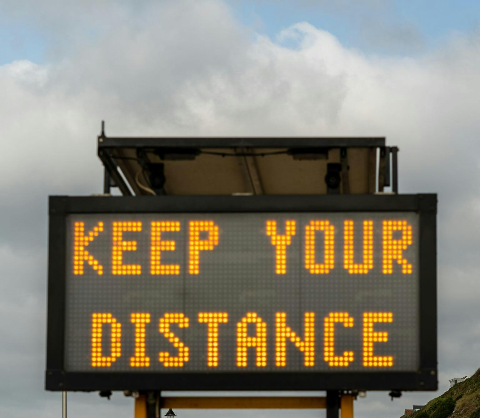 a large LED road sign is pictured in from of blue sky and grey clouds. The sign reads keep your distance in yellow capital letters
