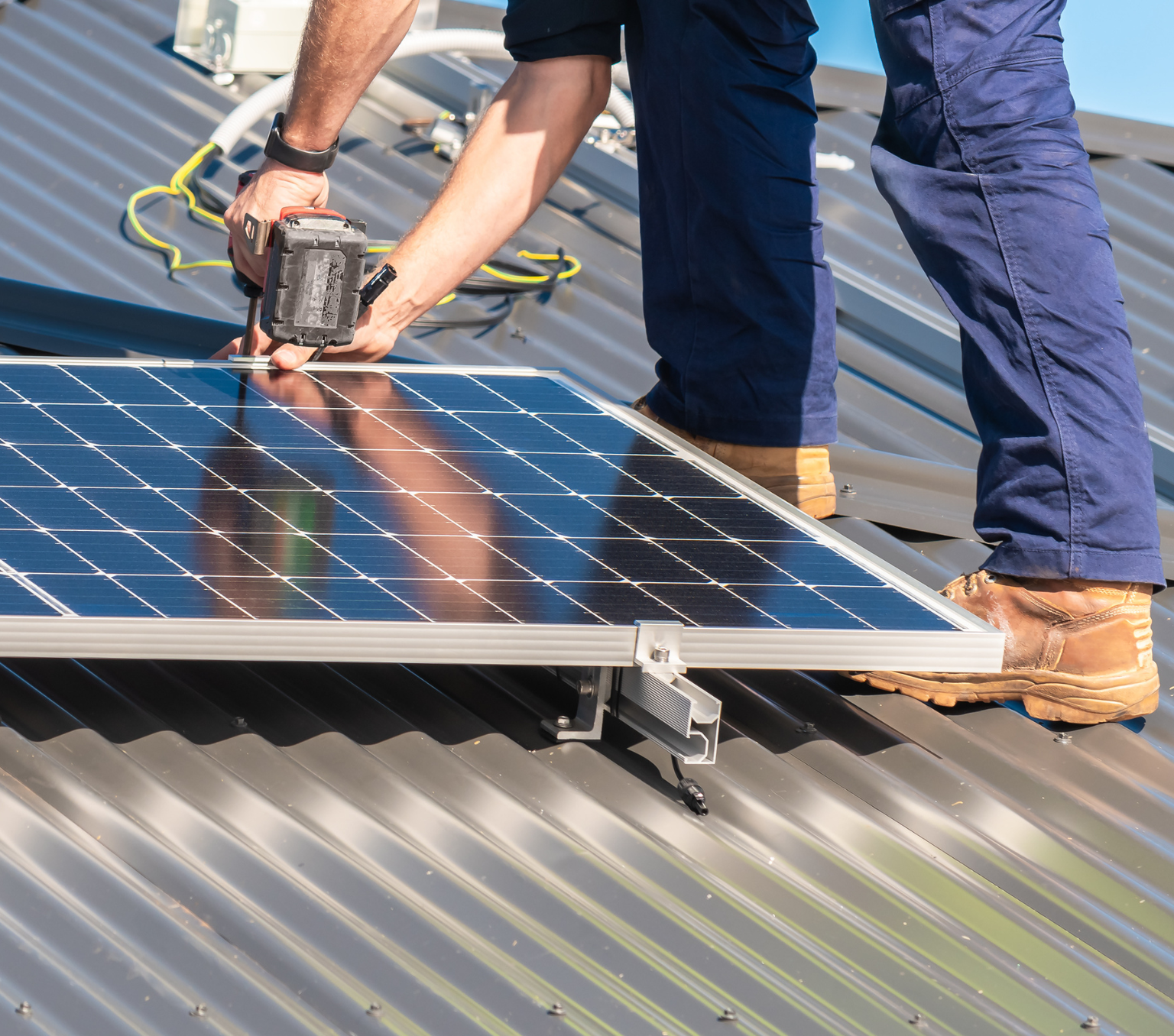 A man stands on a roof, bent over while using a drill near a solar panel.