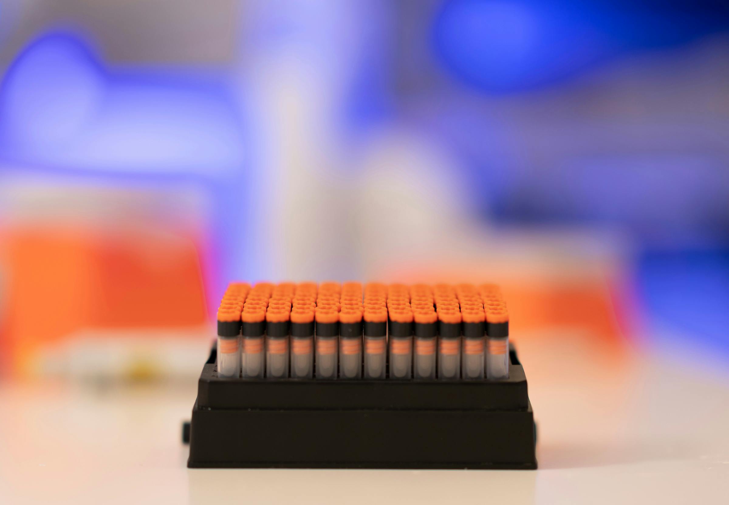 Black tray filled with plastic vials with orange lids in front of a blurred laboratory background