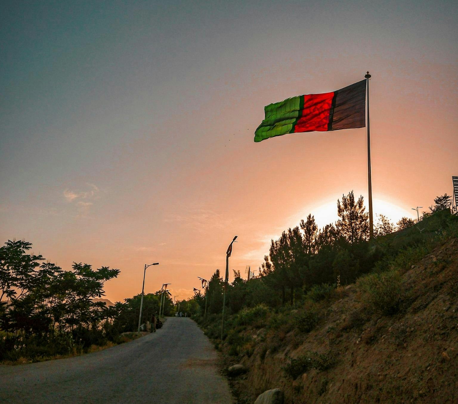 Afganistan flag with black, red and green vertical stipes and a yellow emblem is flying on a flag post on the side of a road. The road is surrounded by shrubs and the sun is setting in the background