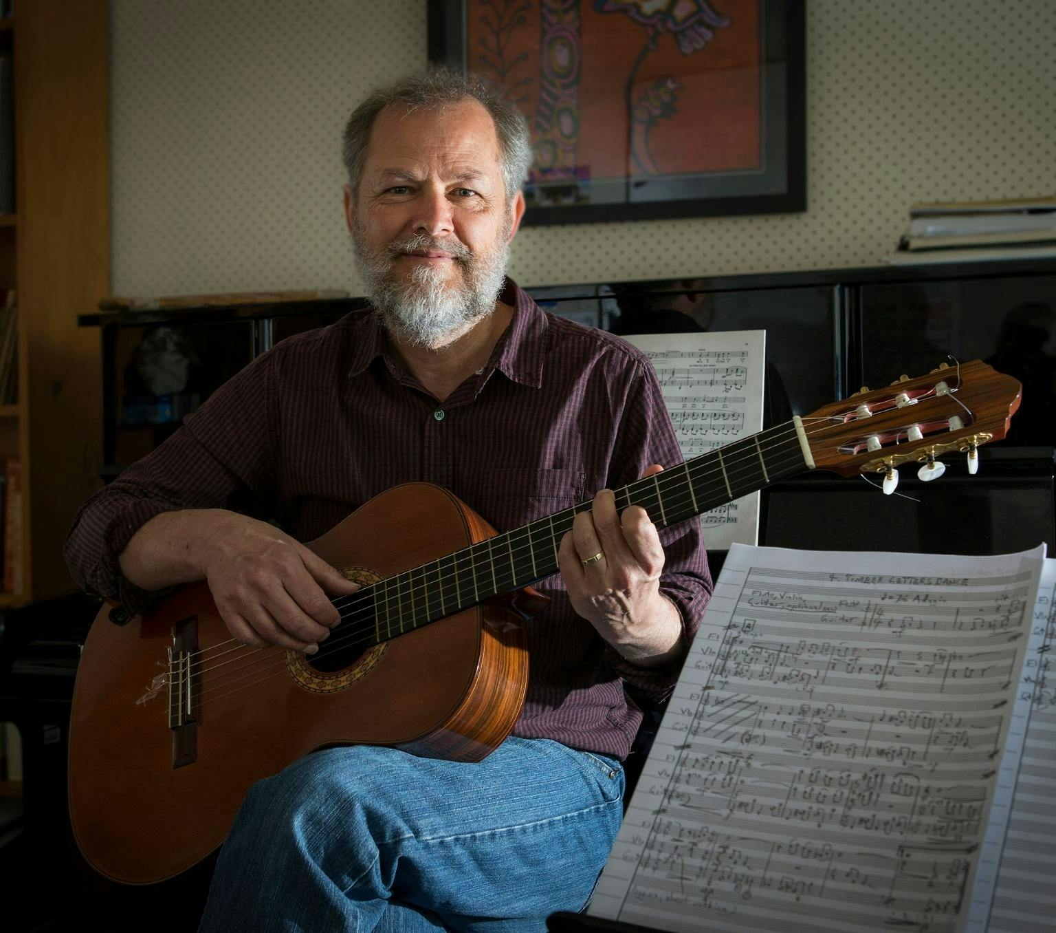Christopher is wearing a maroon collared shirt and blue jeans. He is sitting on a chair and holding a wooden guitare. He is smiling at the camera. next to him is a music stand with music.